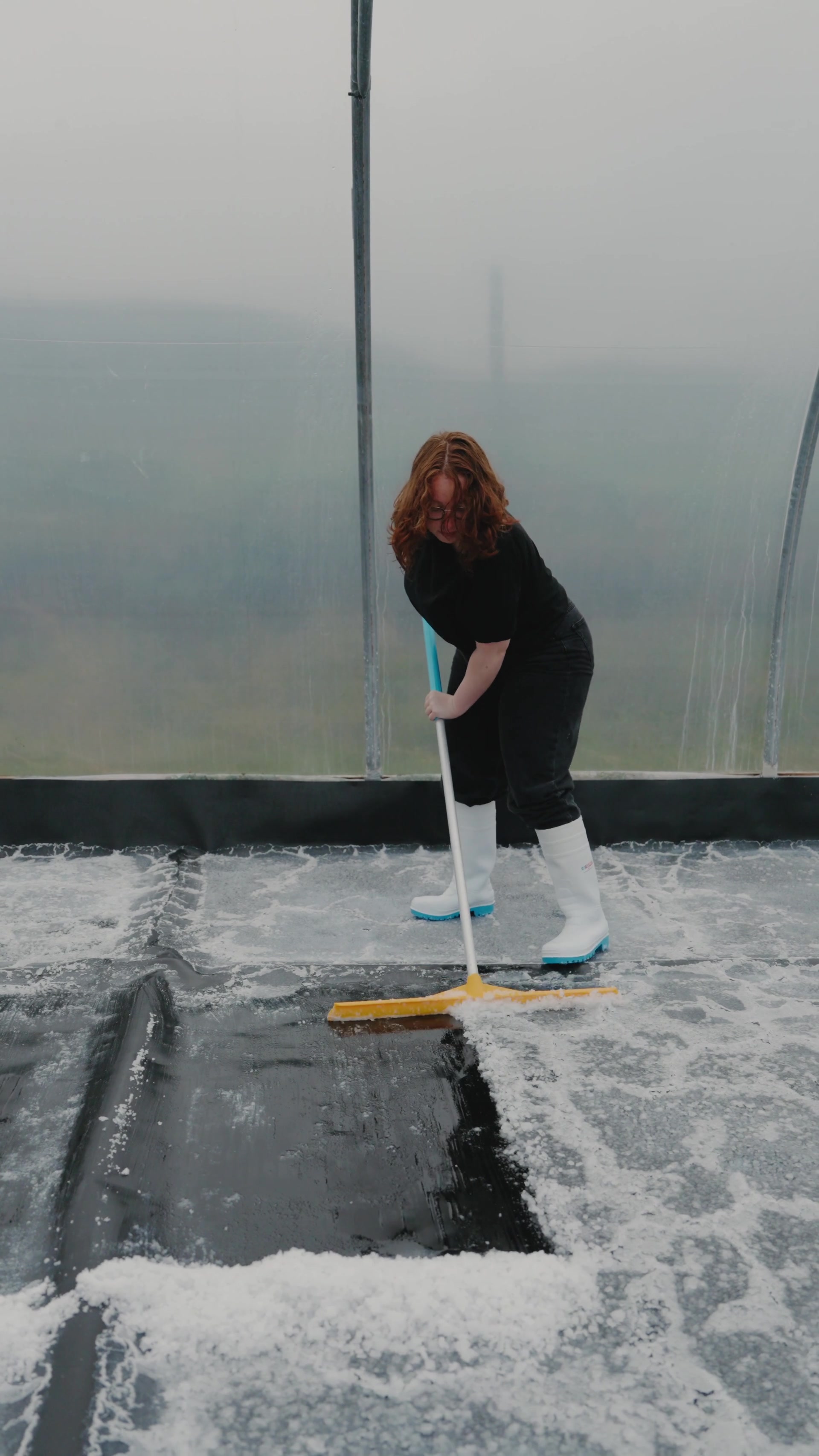 A woman in glasses scrubs a frosted polytunnel floor with a squeegee broom, white wellies planted in the melting ice.