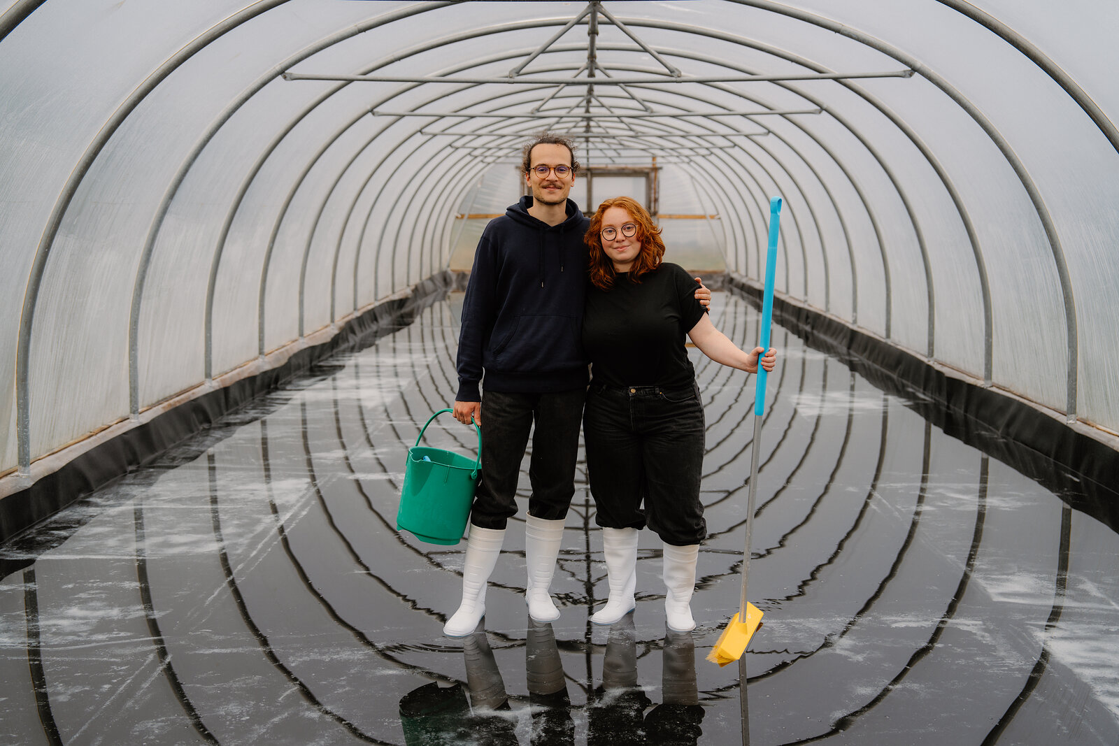 A couple in white boots stand together holding a bucket and squeegee inside a seawater polytunnel at Isle of Skye Sea Salt Company.