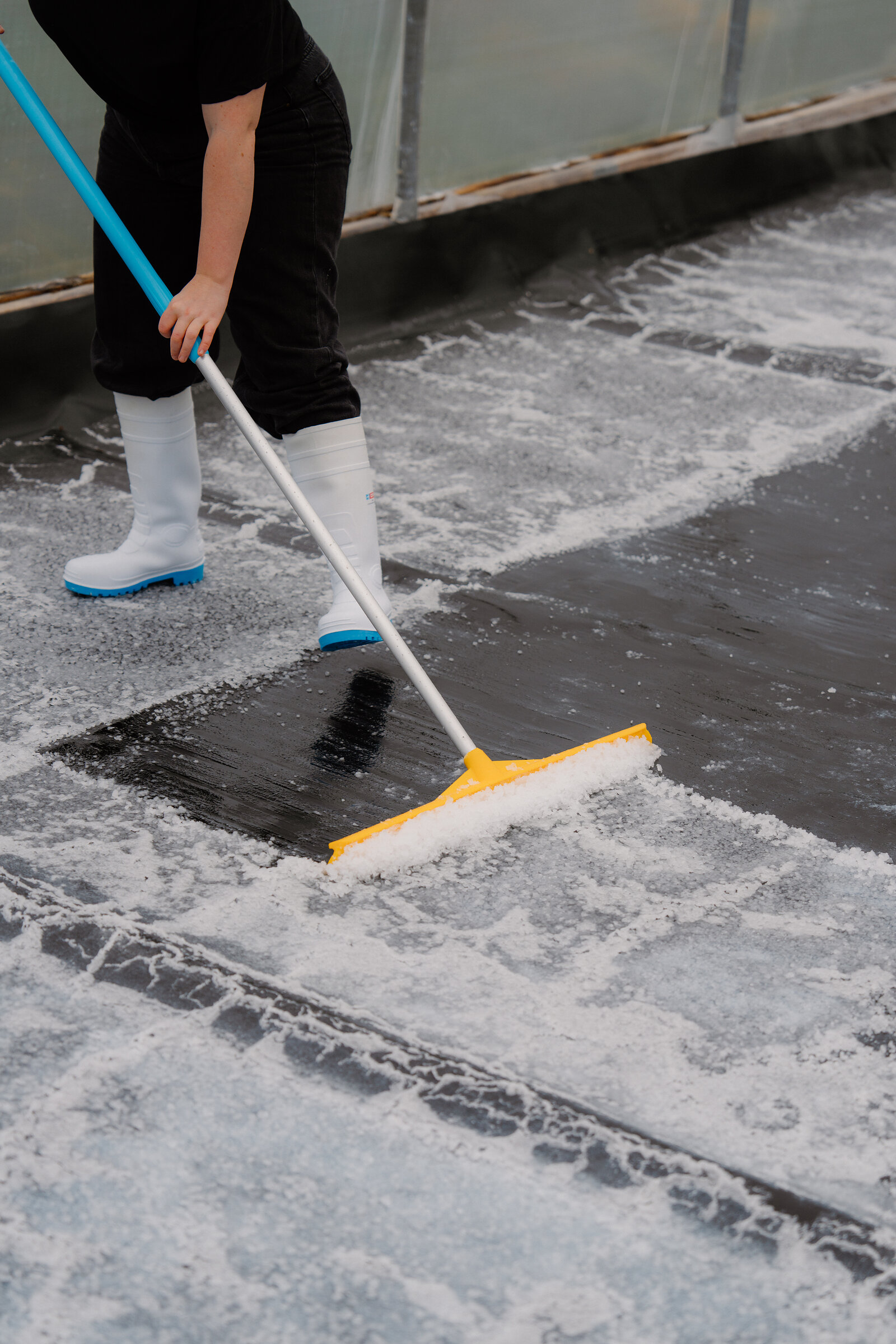 A person in white wellies drags a yellow squeegee across a salt-crusted tarpaulin at Isle of Skye Sea Salt Company.