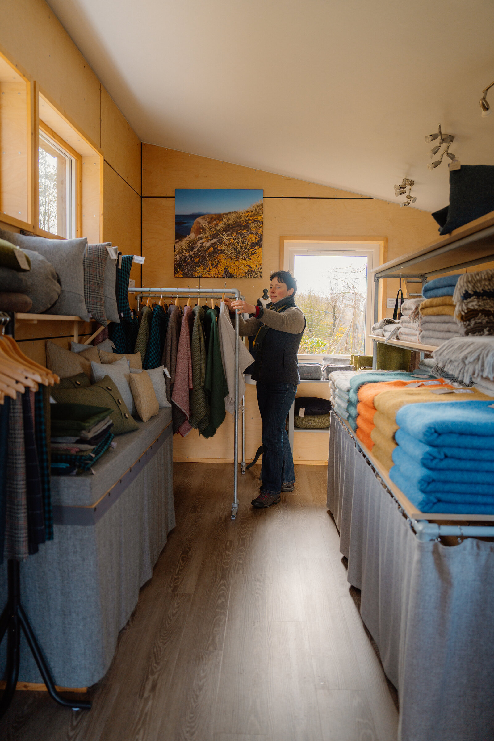 A woman looks through hanging tweed clothing beside shelves of folded throws in a Skye Weavers shop.