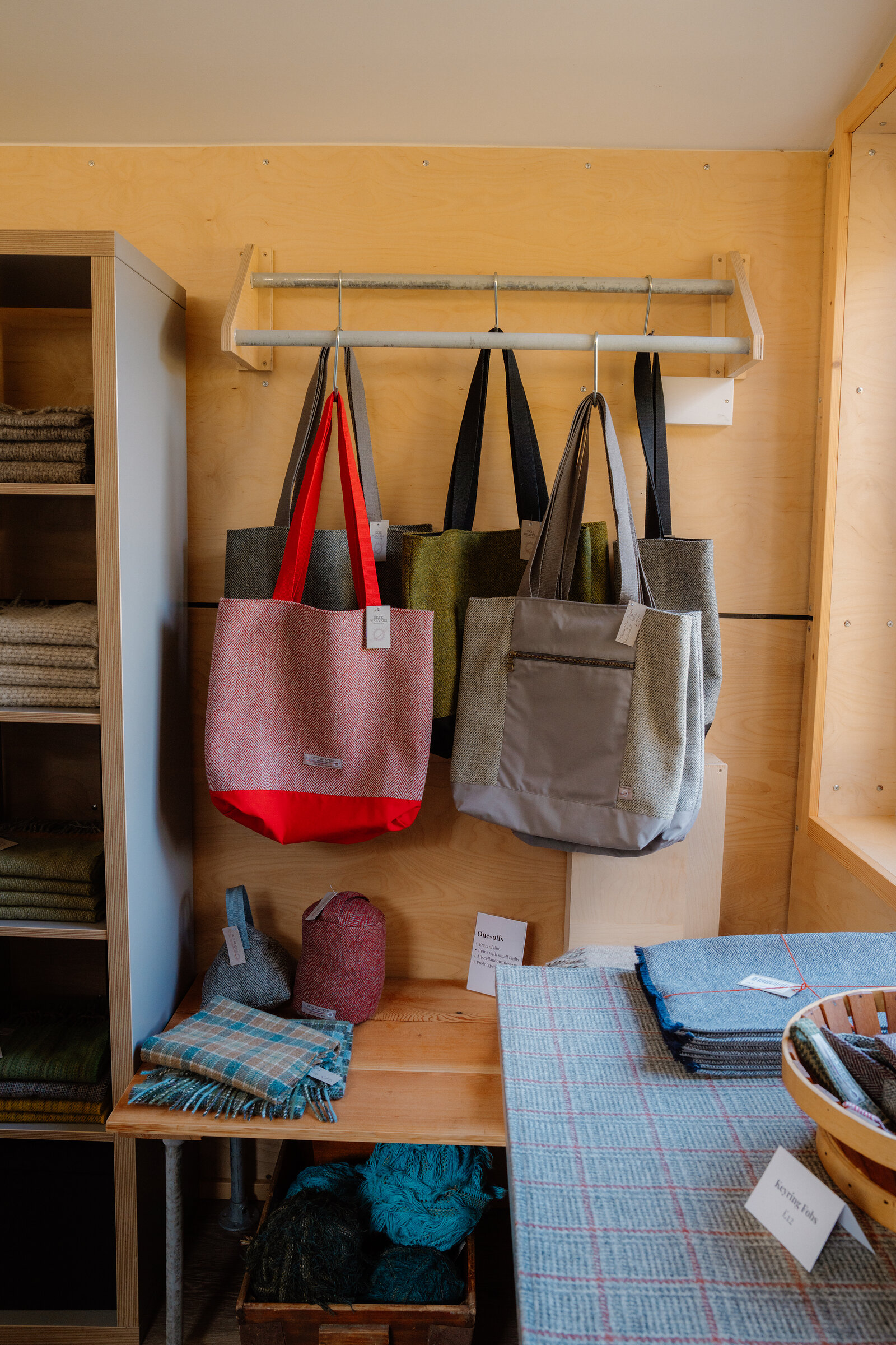 Tweed tote bags in red, olive and grey hang from a rail above folded scarves at Skye Weavers.