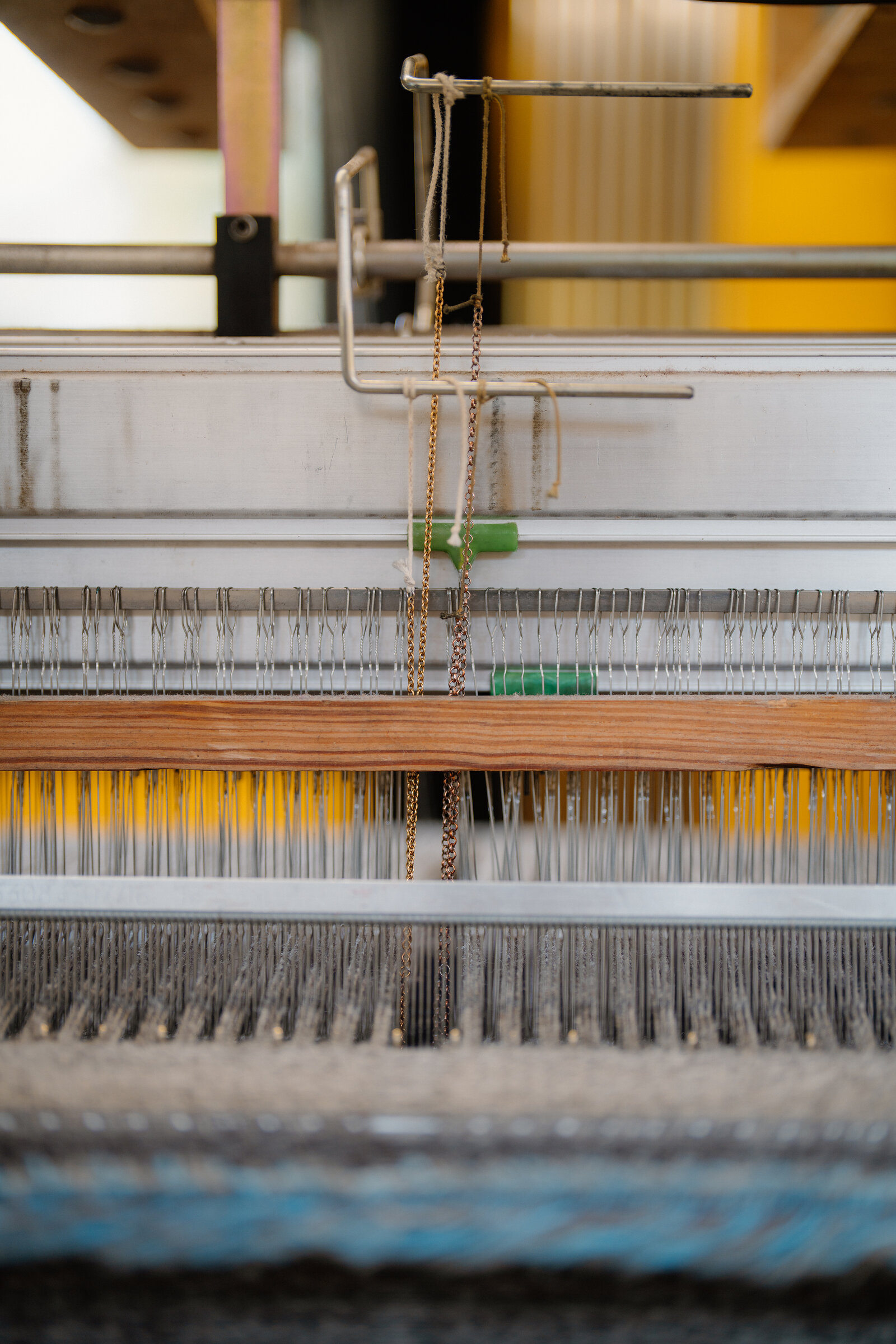 Close-up of a loom's heddles strung with yellow and grey threads beneath a wooden beam