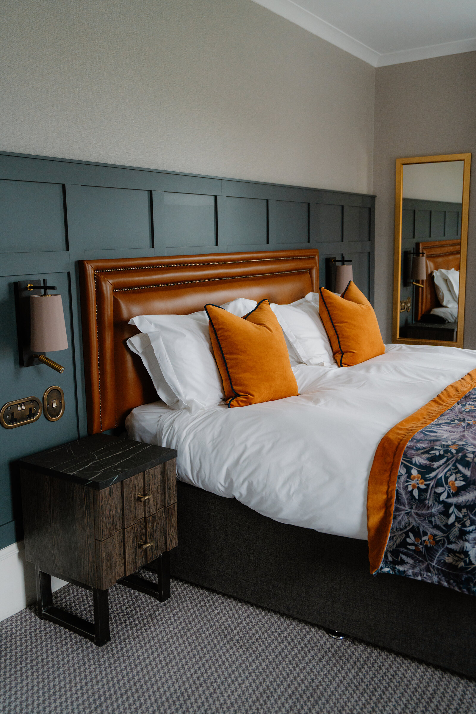 A bed with orange cushions and a leather headboard sits against a teal-panelled wall beside a dark bedside cabinet at Skeabost, part of the Sonas Collection.
