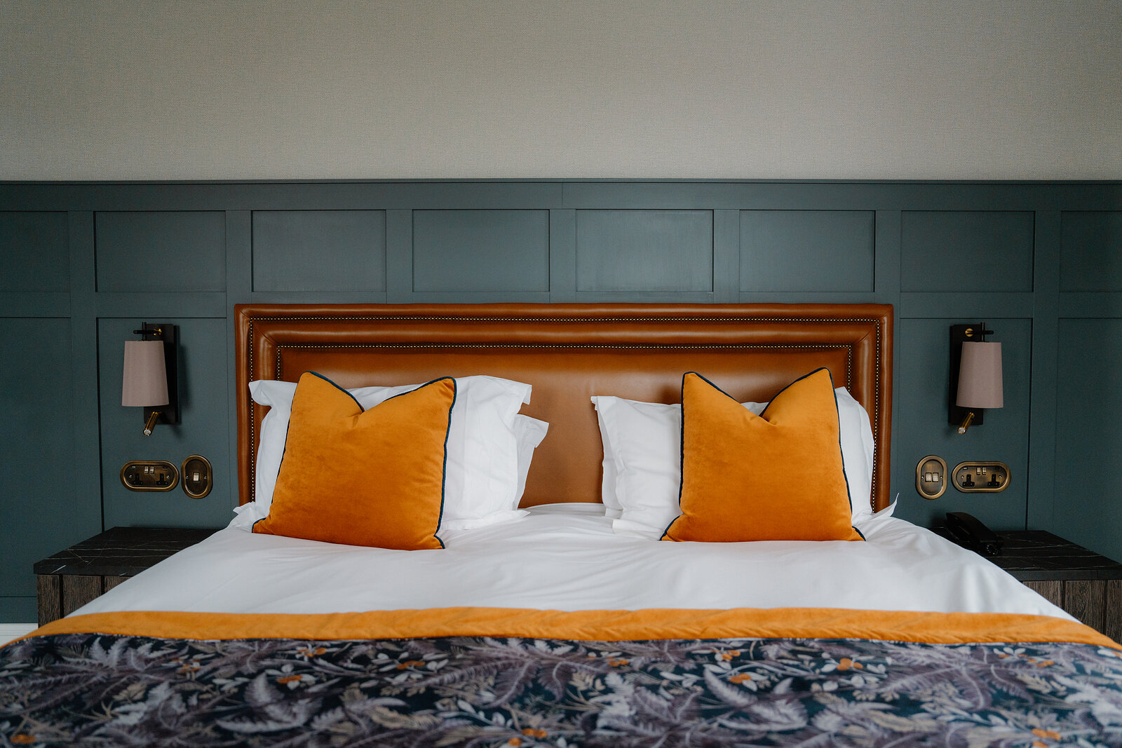 A bed with orange cushions stands against a dark teal tufted headboard flanked by wall lamps in a Skeabost bedroom, part of the Sonas Collection.