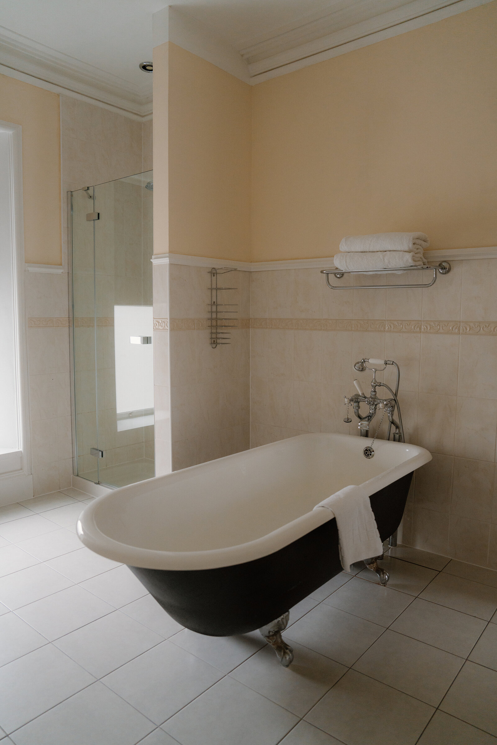 A black clawfoot bathtub stands beside a glass shower cubicle in a cream-tiled Skeabost bathroom, part of the Sonas Collection.