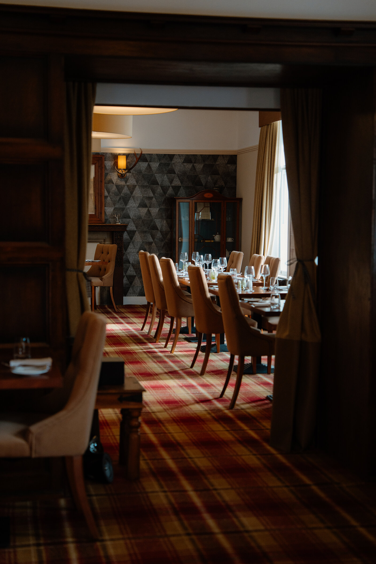 Tables are set on tartan carpet in a wood-panelled dining room at Skeabost, a glass cabinet visible through an archway.