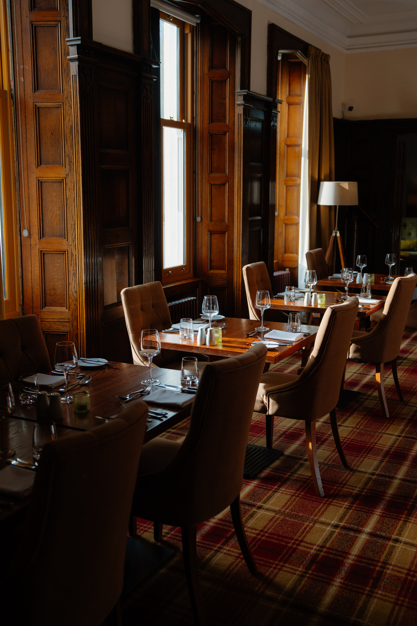Tall wood-panelled windows light a row of set dining tables on tartan carpet at Skeabost.