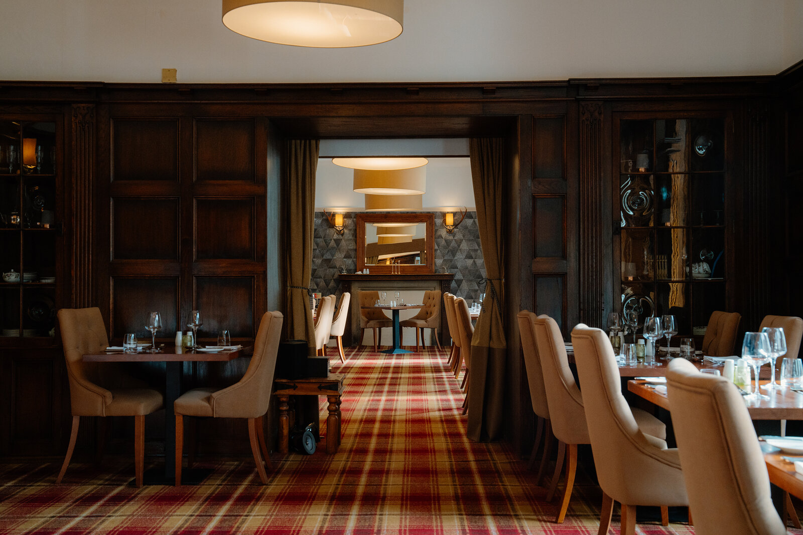 Tables are set on tartan carpet in a wood-panelled dining room at Skeabost, opening to a bar area through an archway.