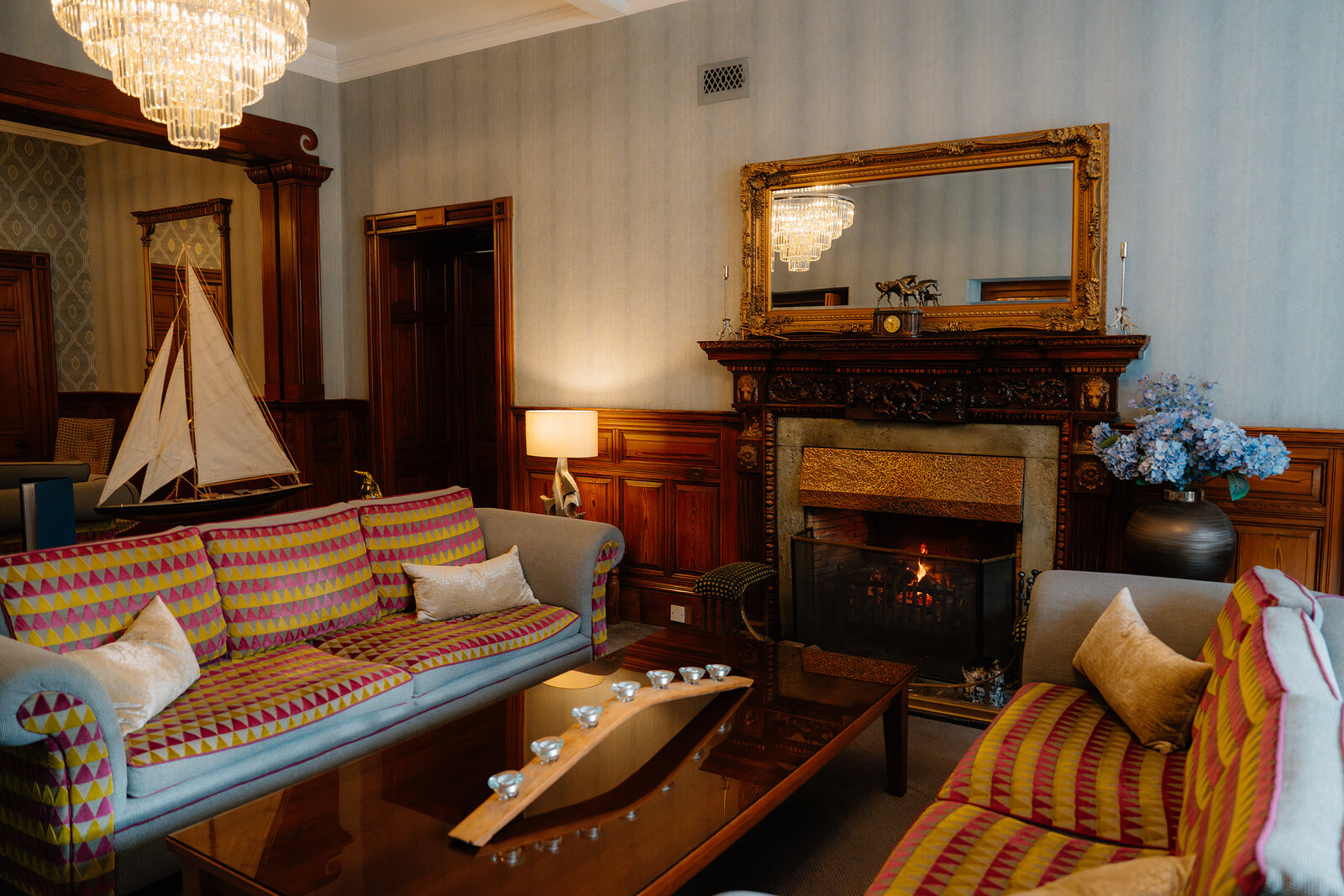 Striped sofas and fresh flowers surround a lit fireplace beneath a gilt mirror in a Skeabost lounge, a model sailboat on the sideboard.
