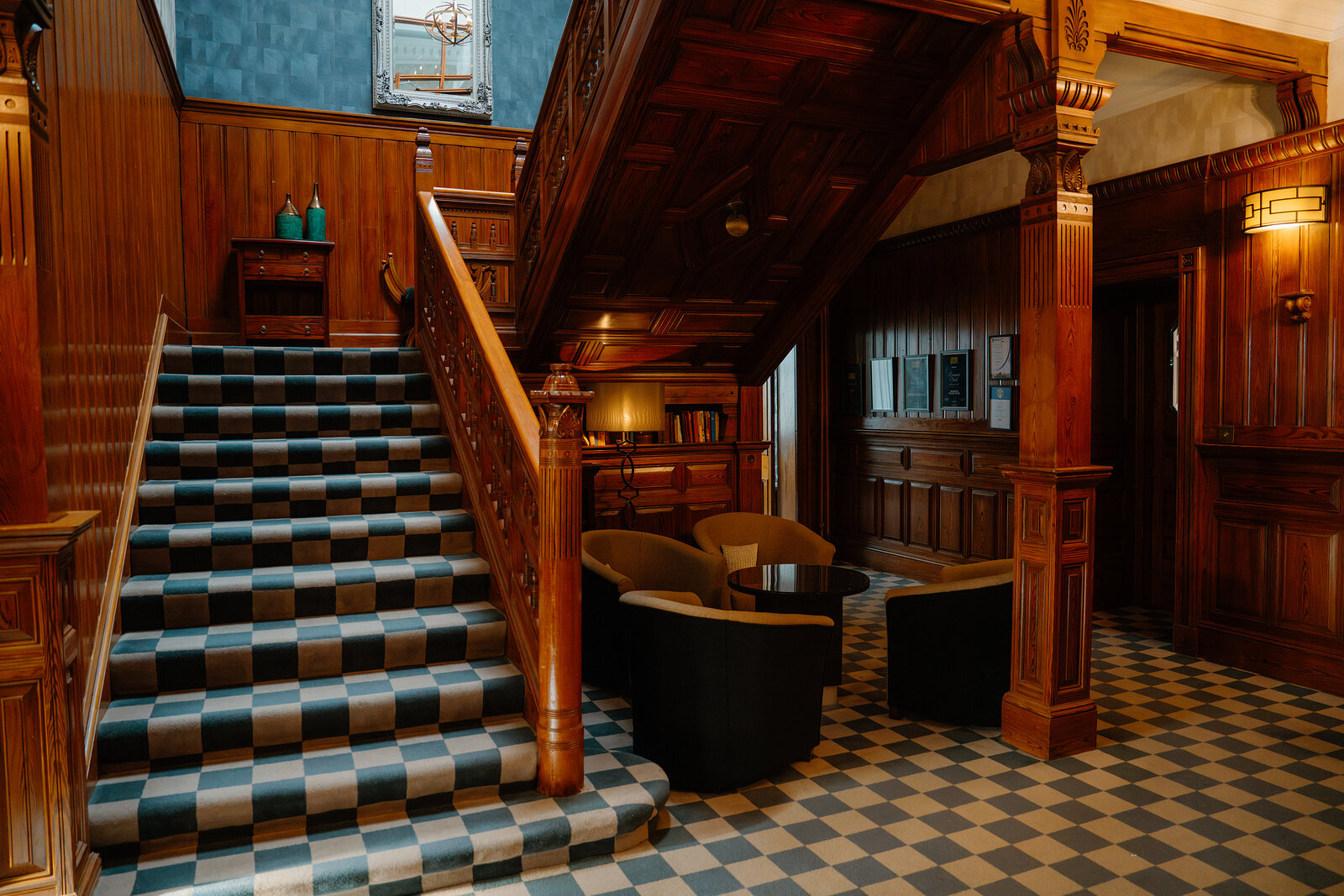 A dark wooden staircase descends beside a checkerboard-tiled floor in Skeabost's entrance hall, armchairs set near a reception desk.