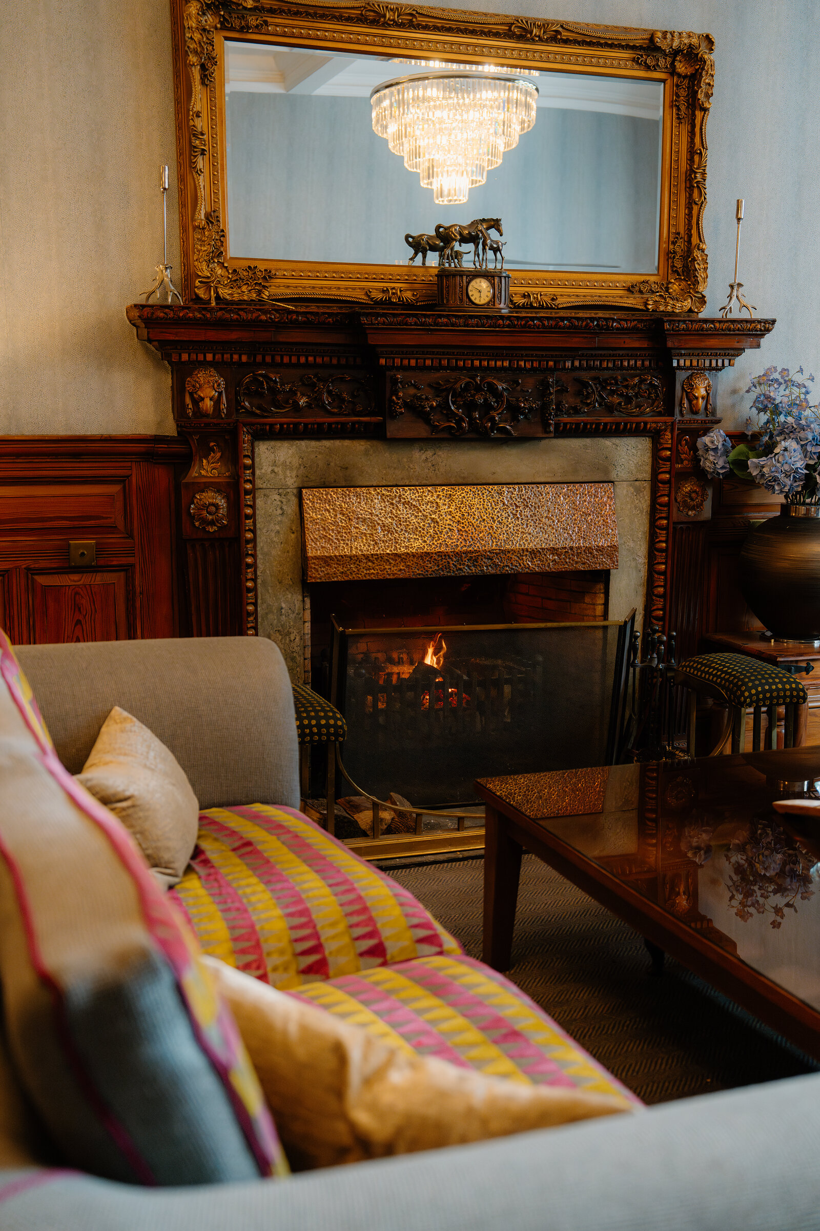 A gilt-framed mirror hangs above a lit fireplace flanked by porcelain ornaments in a striped-sofa Skeabost lounge.
