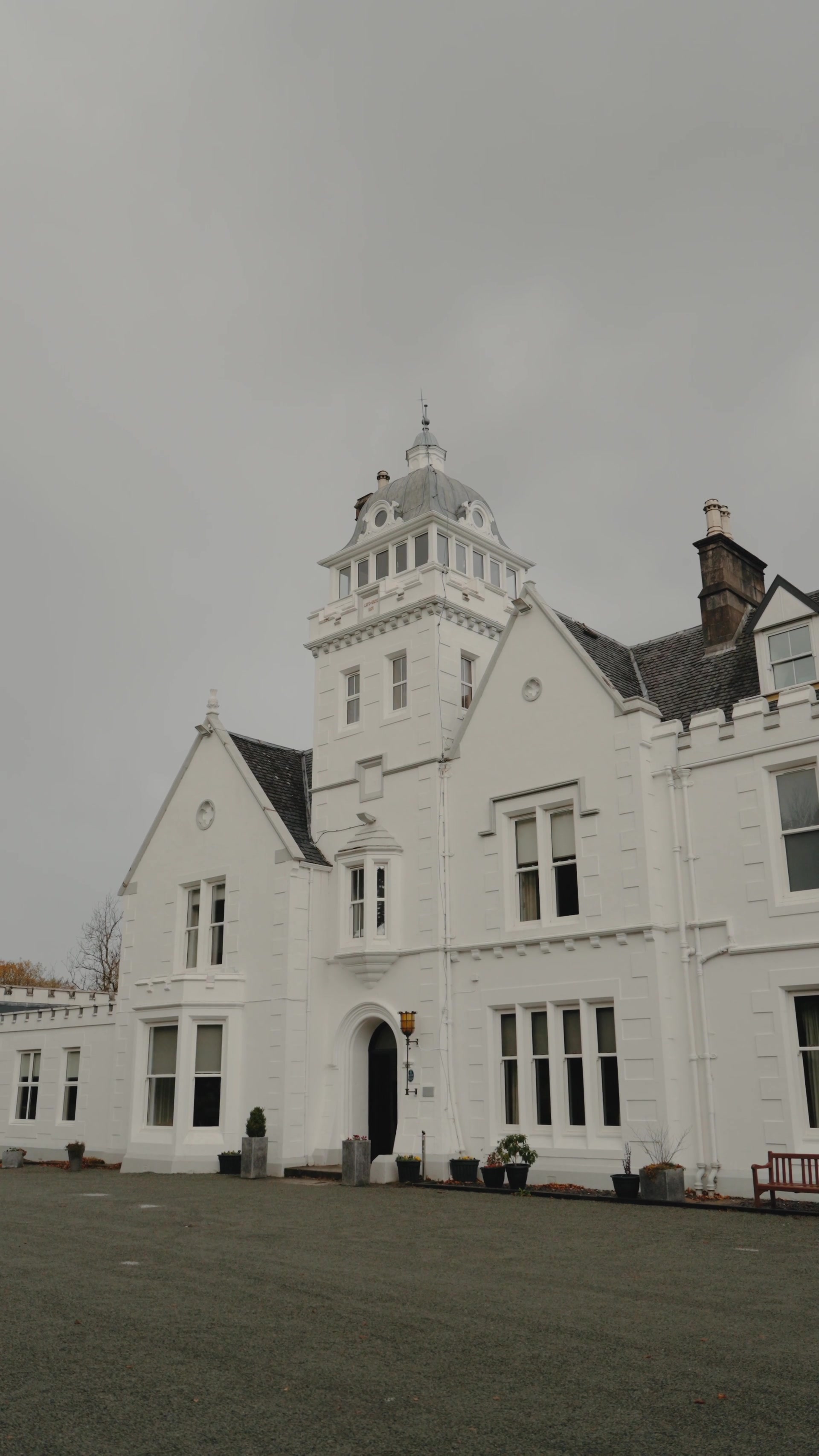 A white Victorian mansion with a domed corner tower and crenellated wings stands beneath a grey sky, potted shrubs lining its entrance.