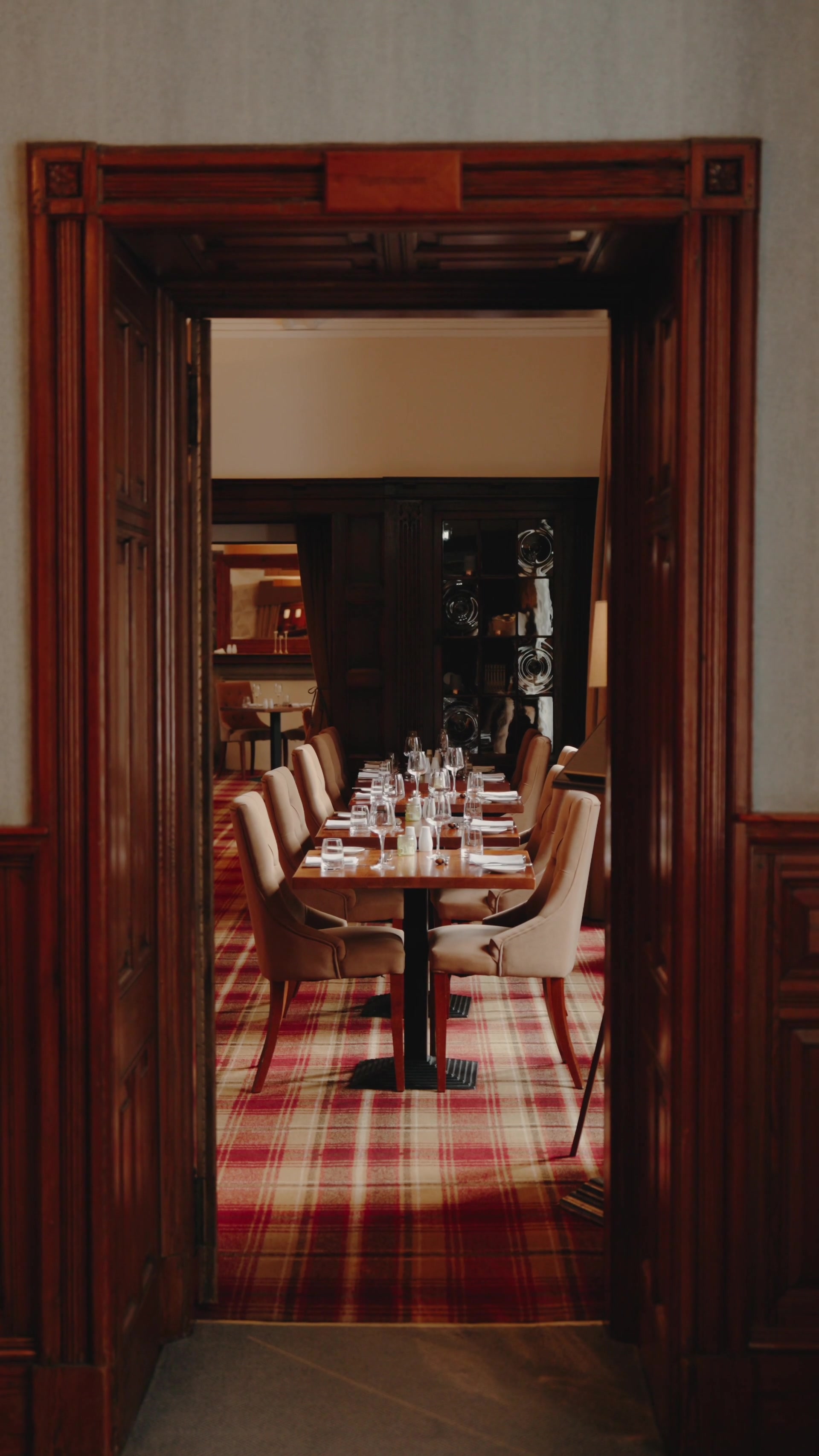A tartan-carpeted dining room set with wine glasses is seen through a carved wooden doorway, a mirrored cabinet of glassware against the far wall.