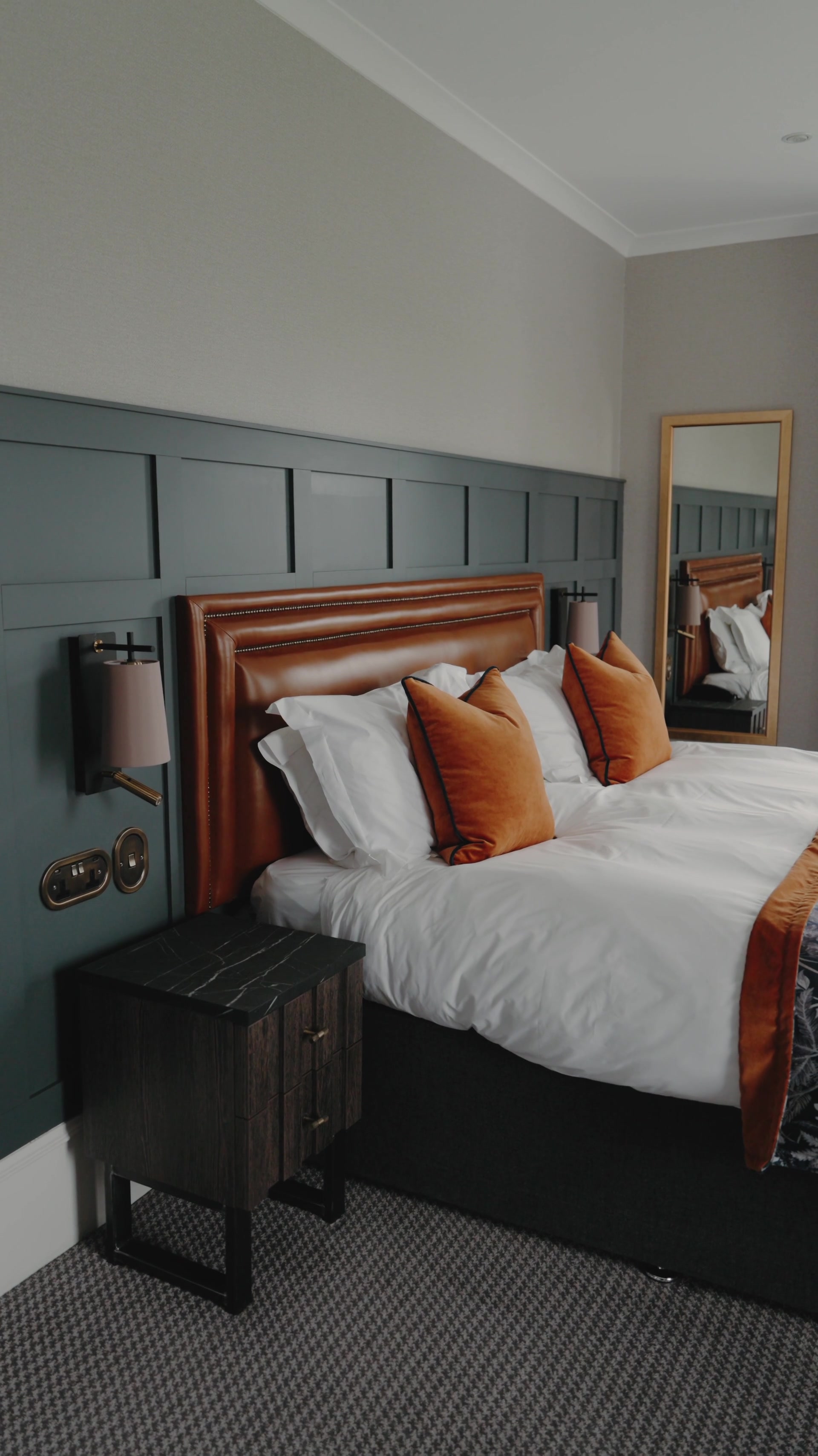 A tan leather headboard is dressed with white pillows and burnt-orange velvet cushions, a gilt mirror reflecting the bed in a green-panelled room.