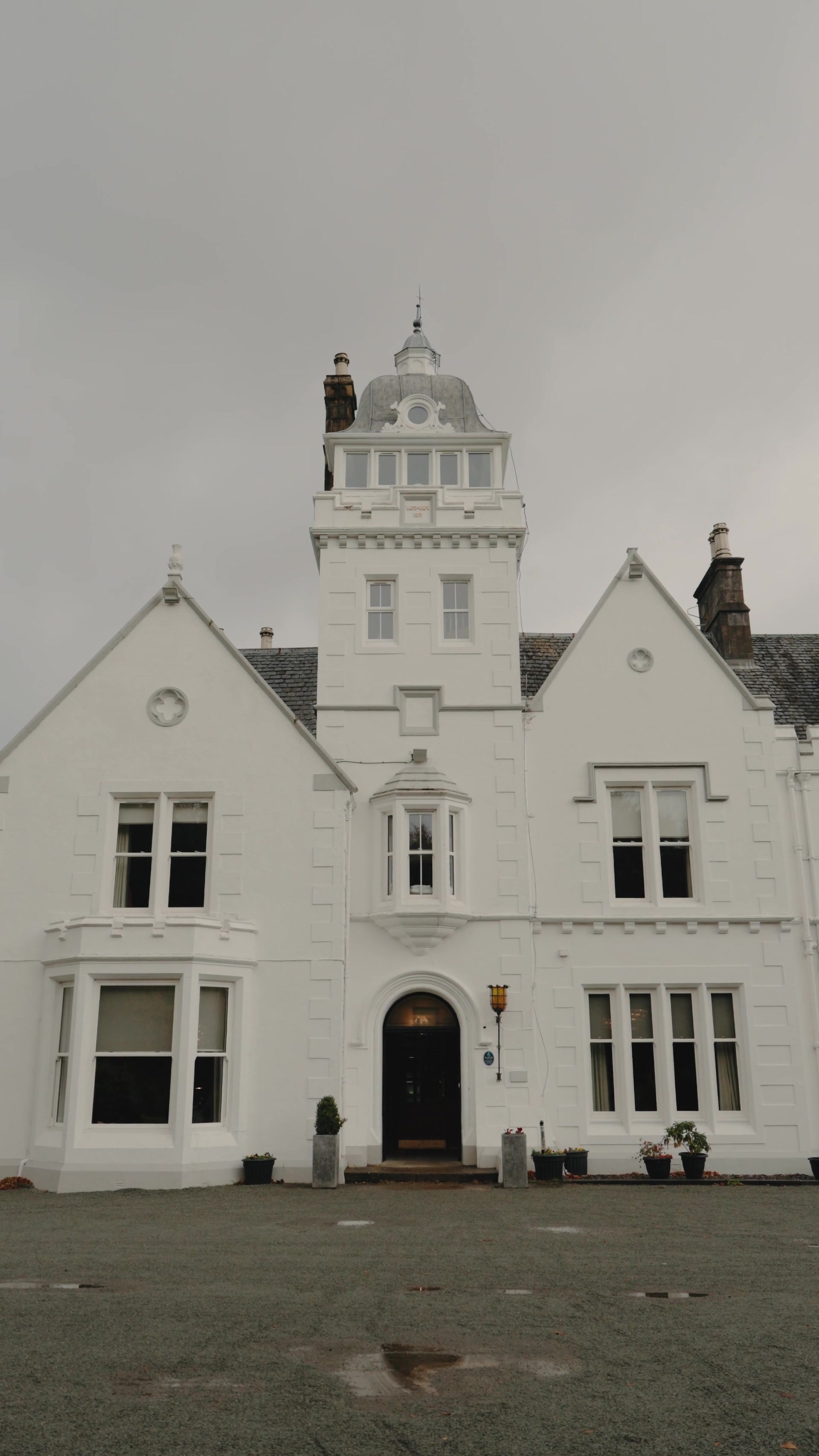 A white Victorian mansion with a domed corner tower and gabled wings rises beneath a grey sky, potted shrubs flanking its arched entrance.