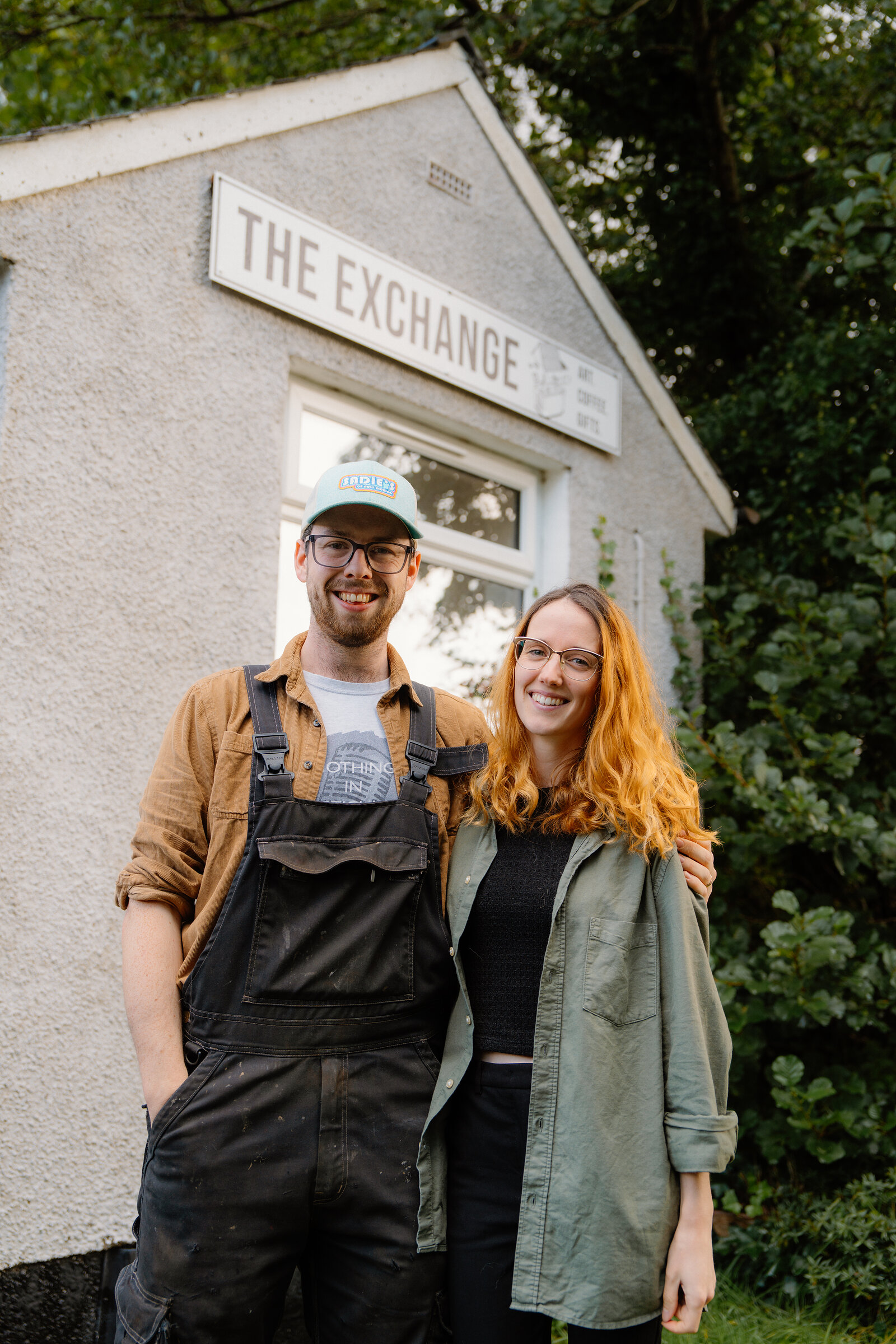 A man in dungarees and a woman stand arm in arm beneath 'The Exchange' sign on Raasay.