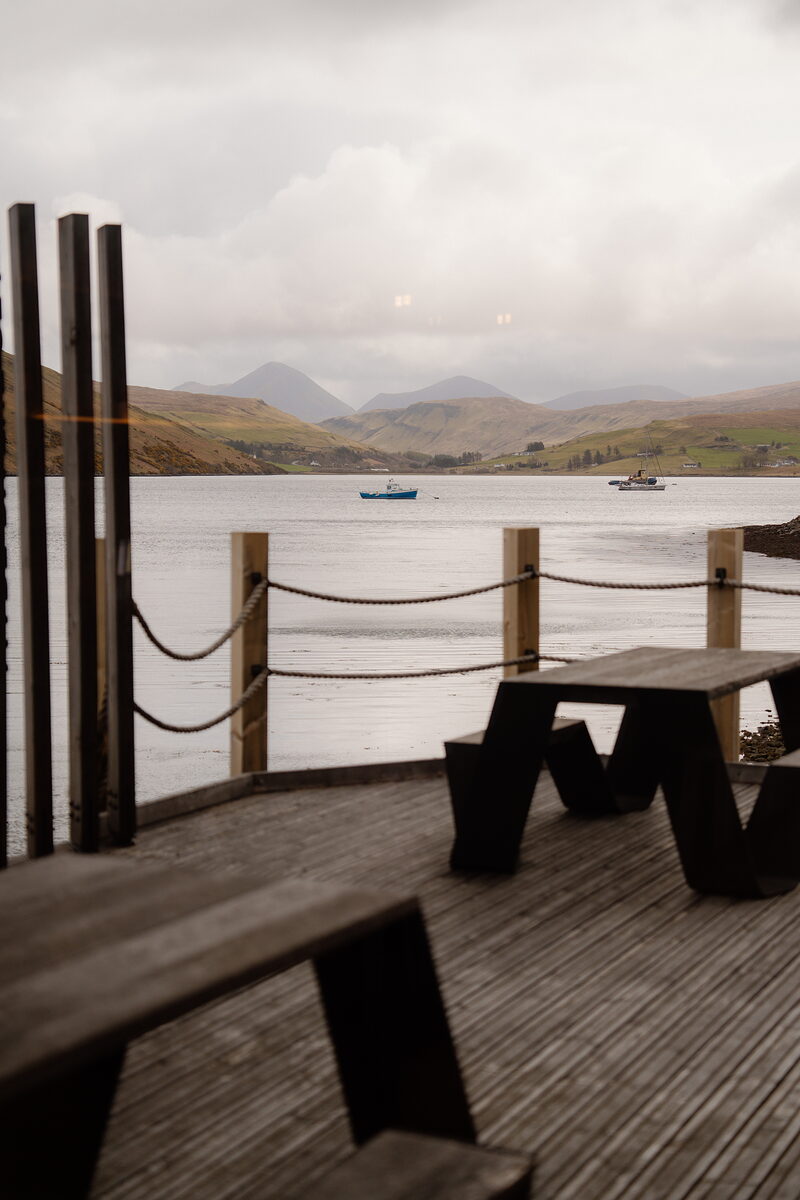 Wooden picnic benches sit on a deck overlooking a sea loch and mountains at Three Chimneys at Talisker, a rope railing along the edge.