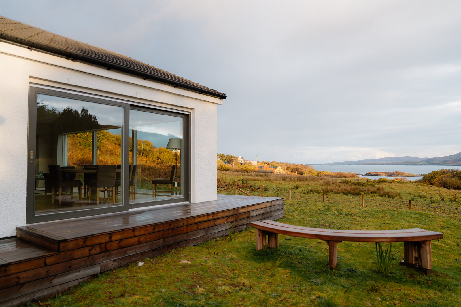 A house with glazed sliding doors opening onto a wooden deck and bench, a loch and hills visible beyond