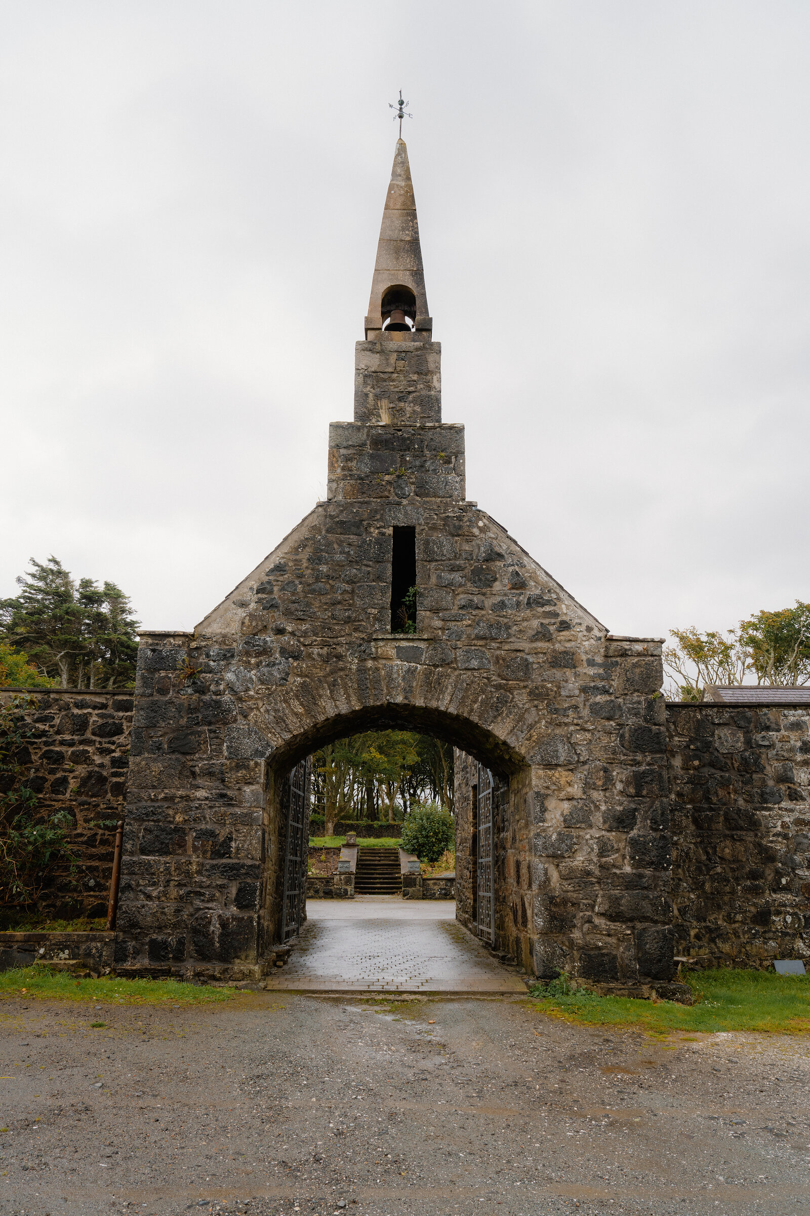 A stone archway topped by a bell tower and weathervane, a gravel path leading through to a garden beyond