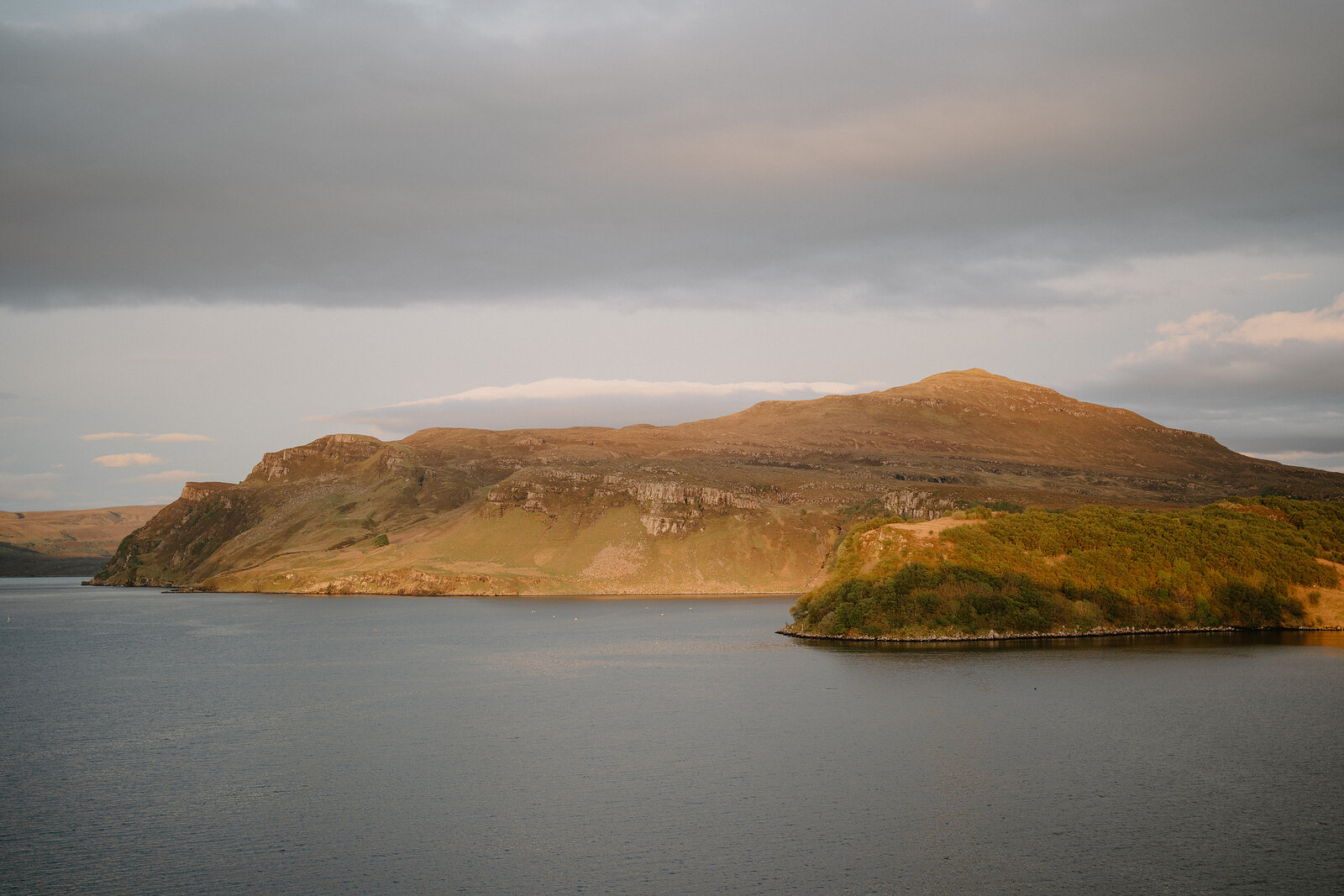 Evening light warms a grassy headland beside a calm loch on the Isle of Skye.