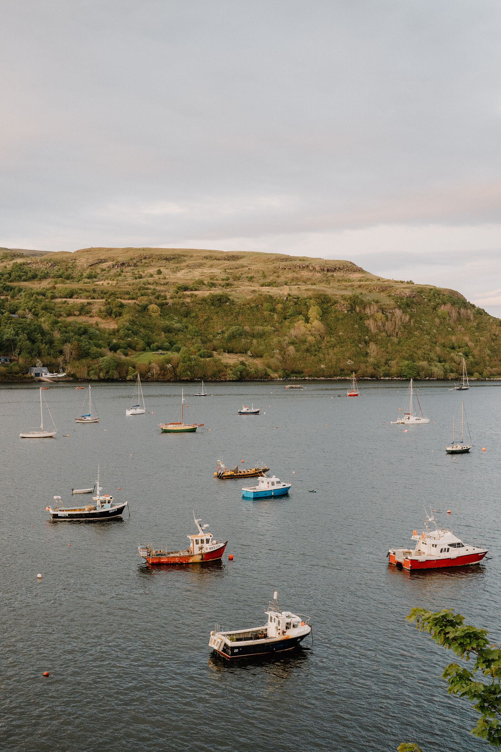 Sailboats and small motorboats lie moored in a calm bay beneath a green hillside on the Isle of Skye.