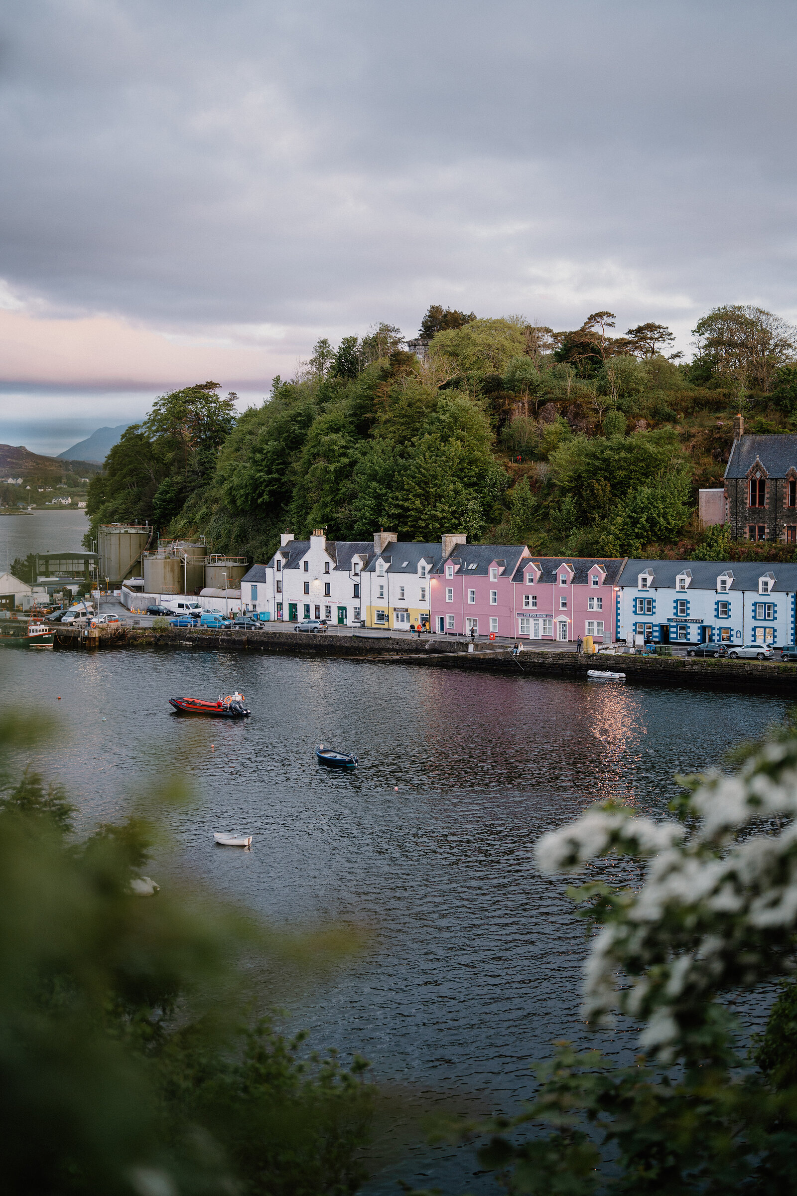 Portree's pink, white and blue harbourfront houses reflect in the water at dusk, boats moored below.