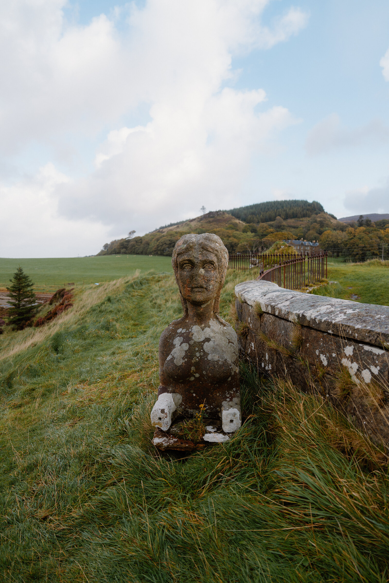 A weathered stone bust of a figure stands beside a low wall, a house visible on the hillside beyond.