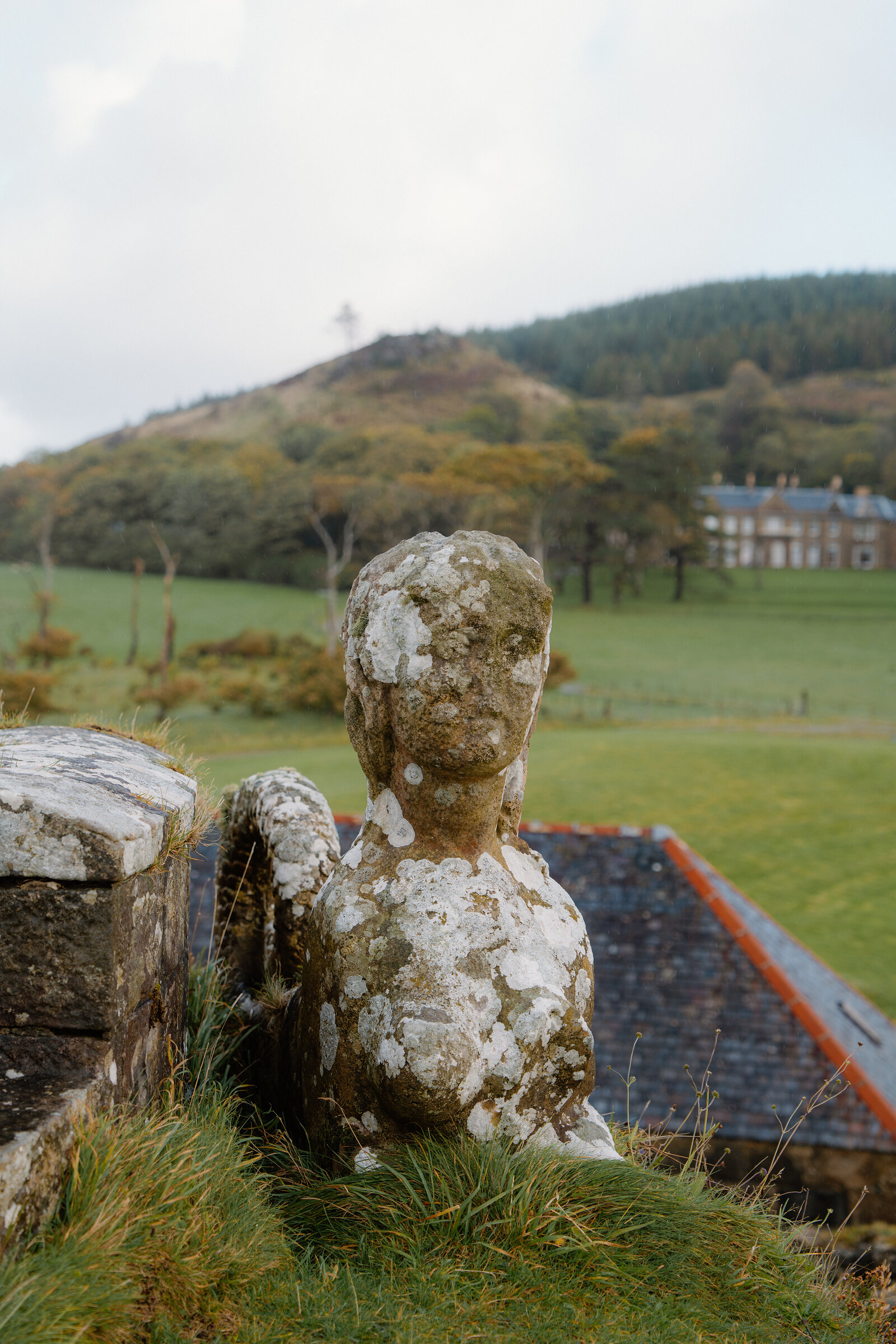A weathered stone bust of a figure sits on a wall before a grand mansion set among green fields.