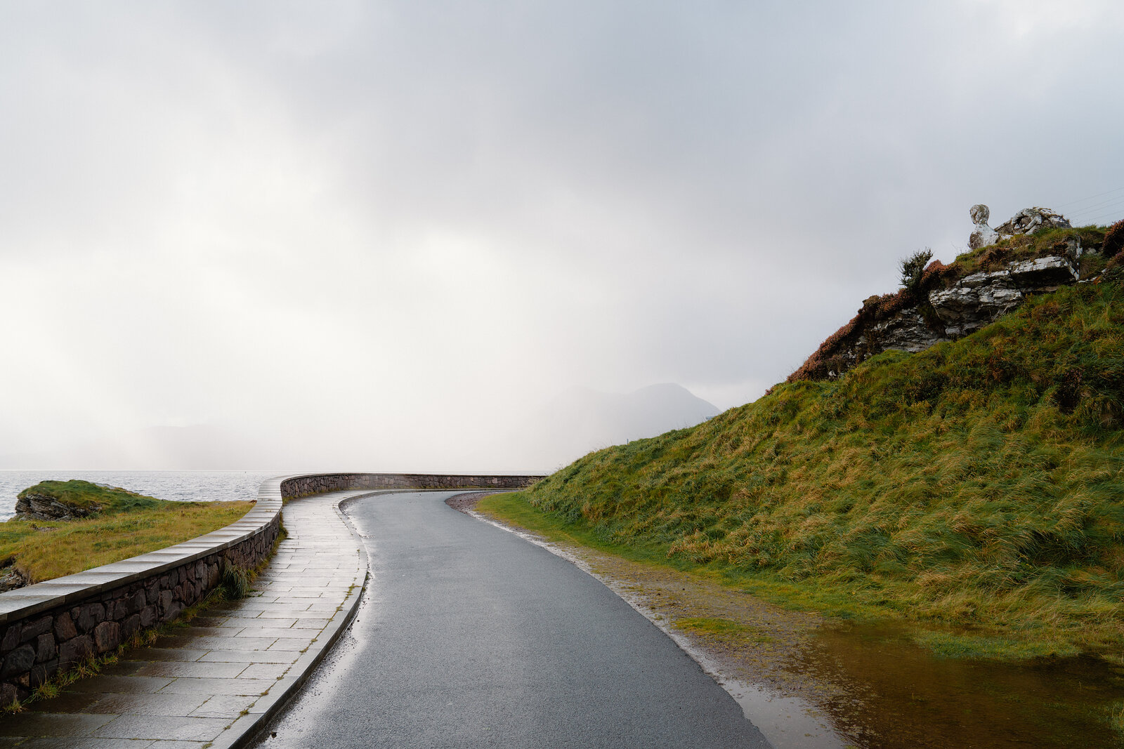 A wet road curves past a low stone wall through mist and grassy slopes.
