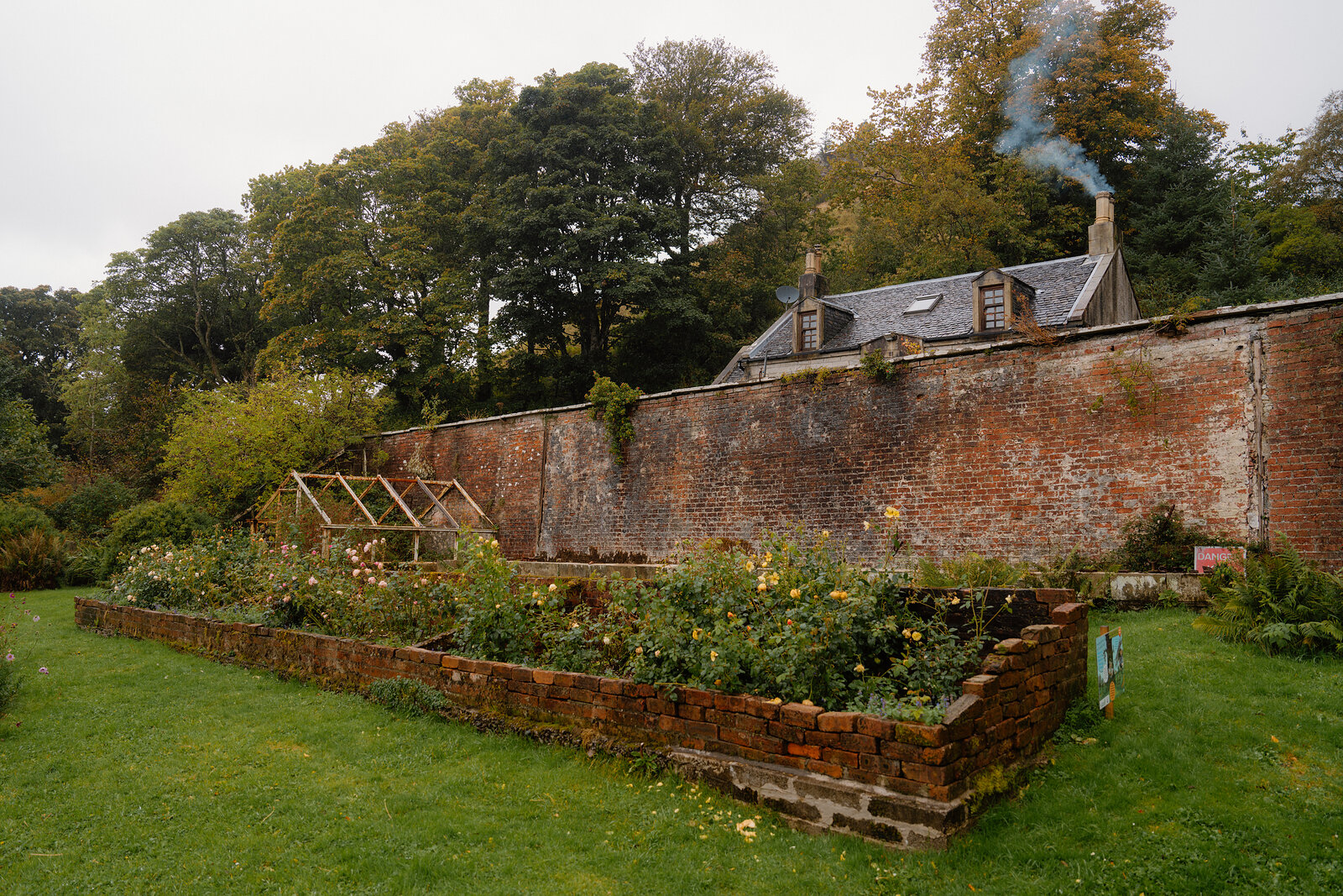 Raised brick flowerbeds and a small greenhouse sit against a walled garden with a cottage chimney smoking beyond.