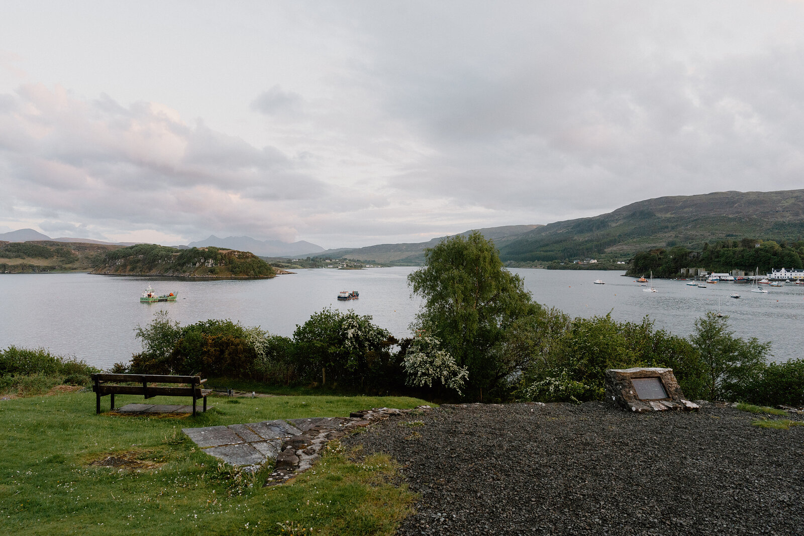 A wooden bench overlooks a loch dotted with small boats beneath wooded hills.