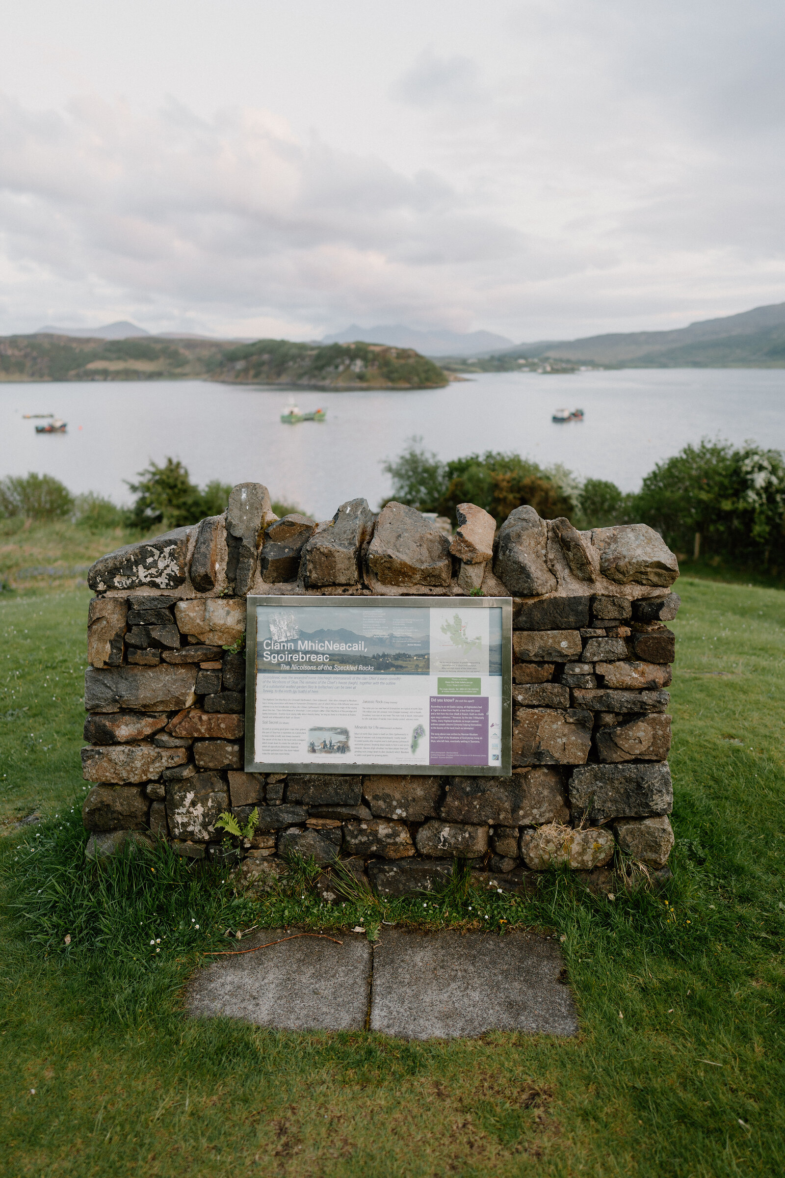 An information plaque set in a stone wall overlooks a loch with moored boats and distant hills.