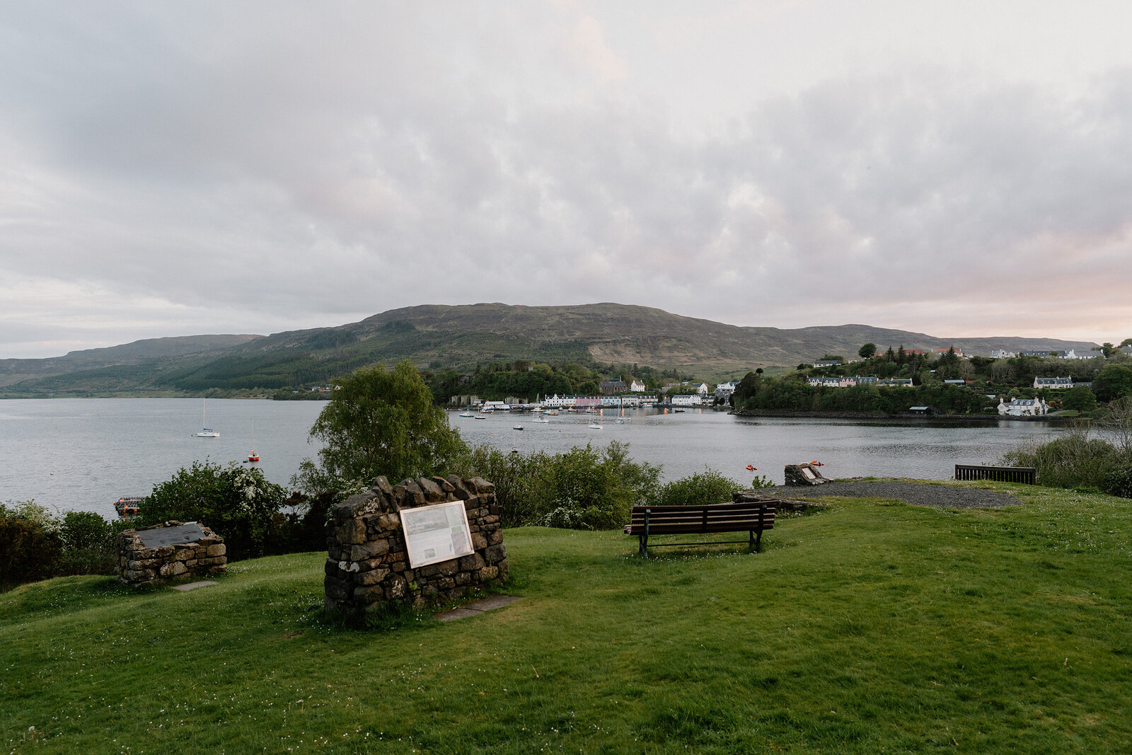 A stone memorial and wooden bench overlook a loch with a village visible on the far shore.