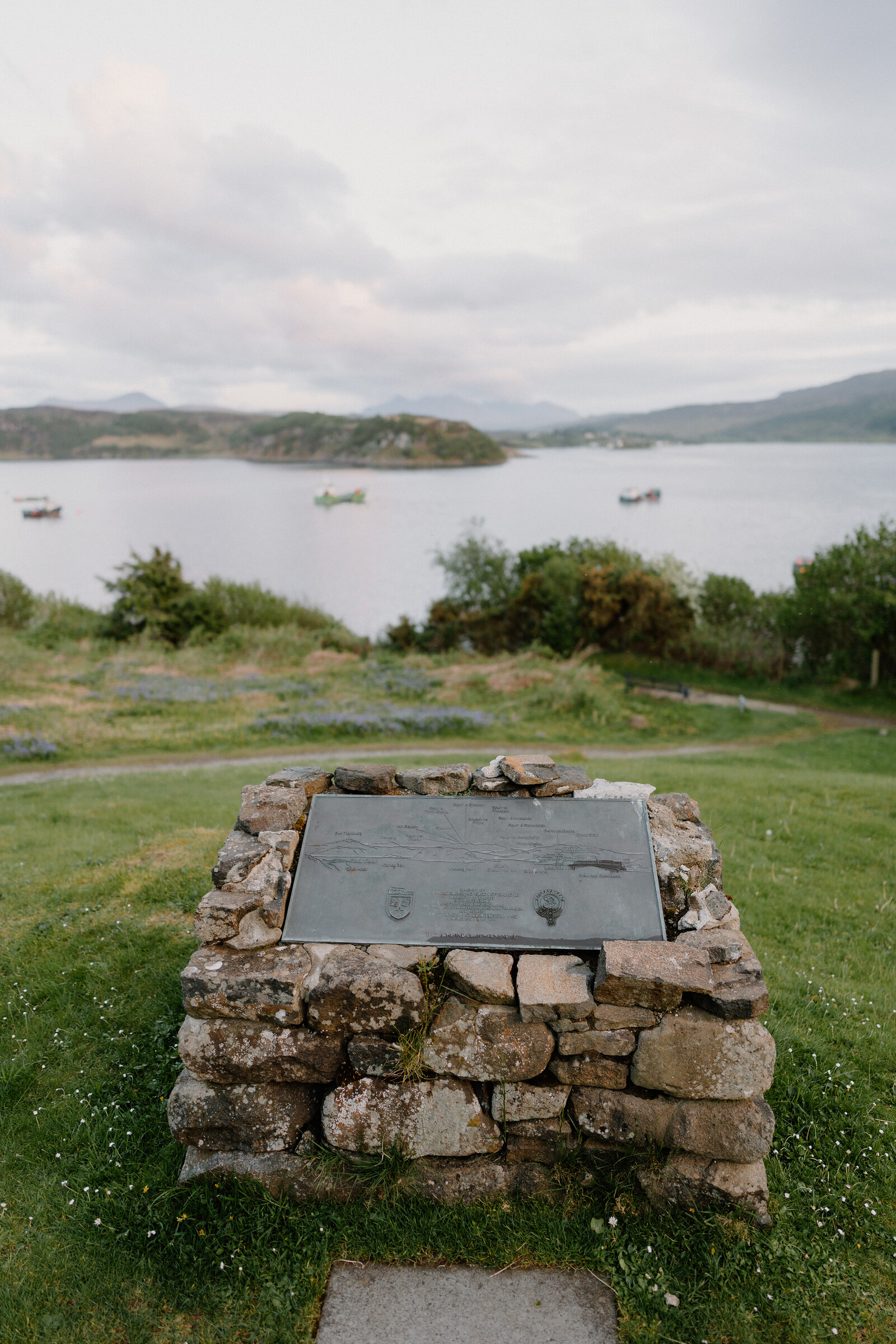 A stone memorial cairn overlooks a bay dotted with small boats.
