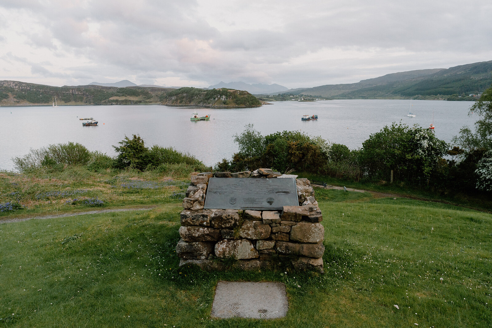 A weathered plaque set in a stone cairn overlooks a calm bay with moored boats.