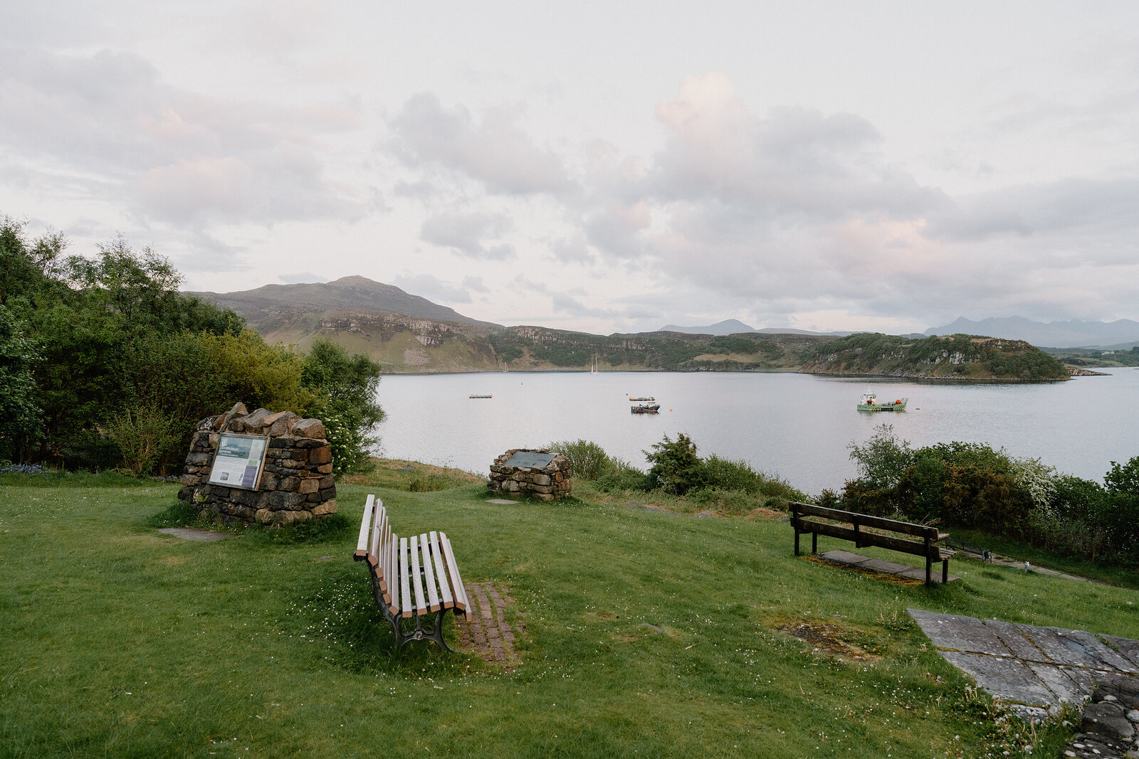 Wooden benches and a stone information cairn sit on a grassy shore overlooking a bay.