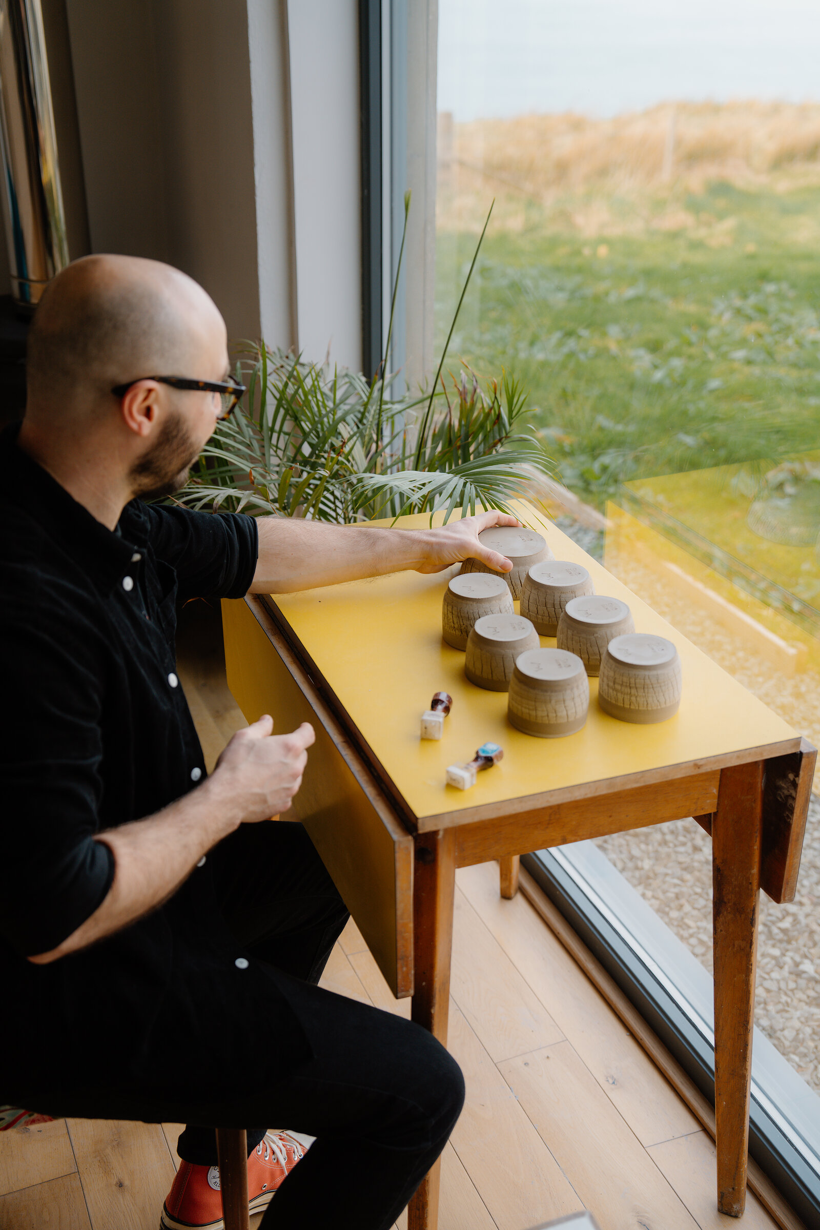 A man reaches toward rows of small ceramic pots lined up on a yellow table by a window overlooking grass and sea.