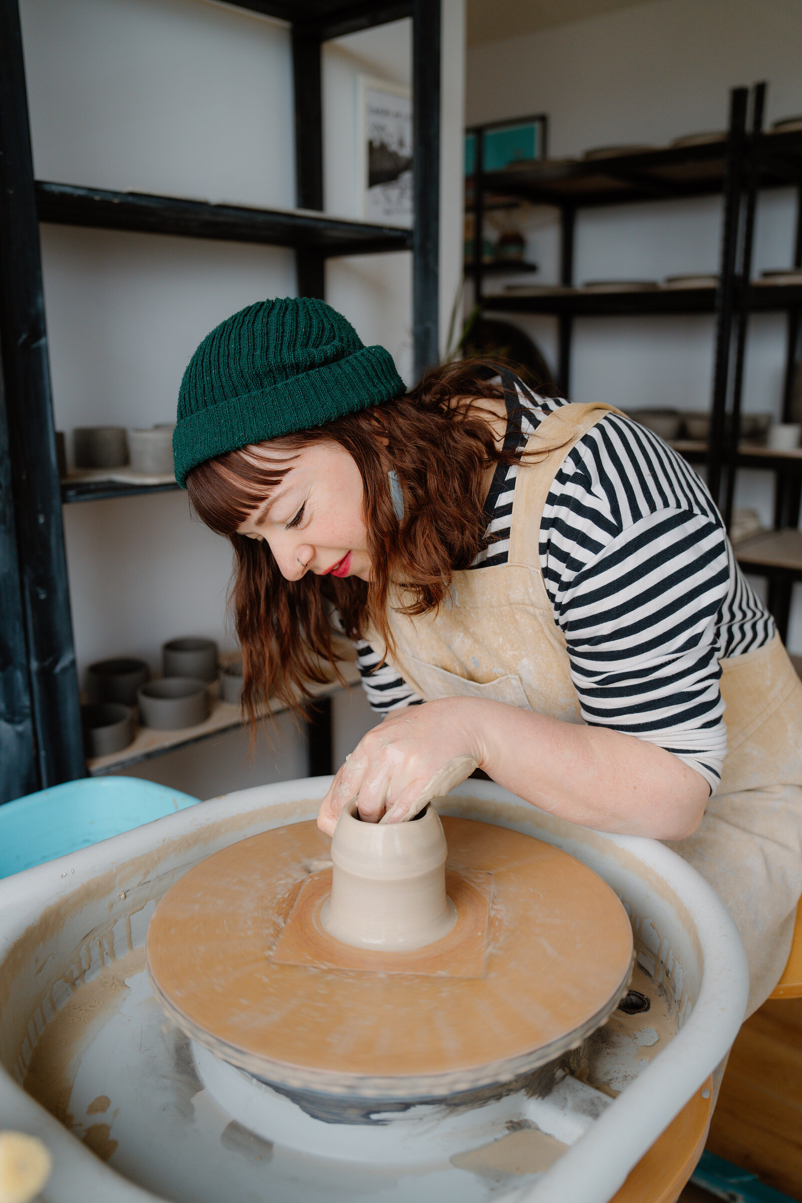 A woman in a green beanie shapes a clay vessel rising on a pottery wheel.