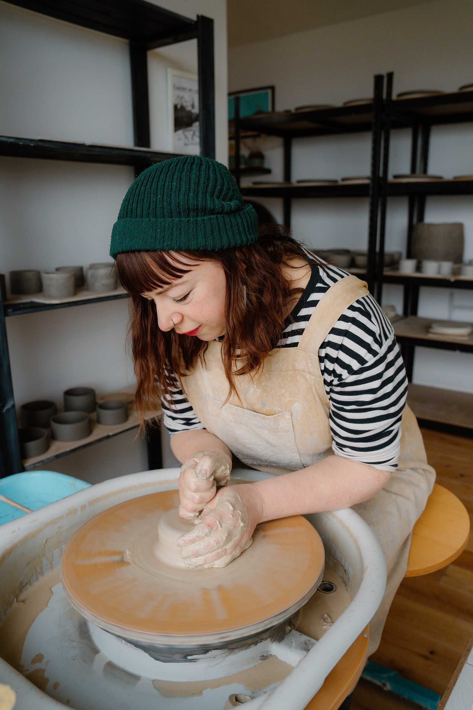 A woman in a green beanie centres a lump of clay on a pottery wheel beside shelves of finished pots.