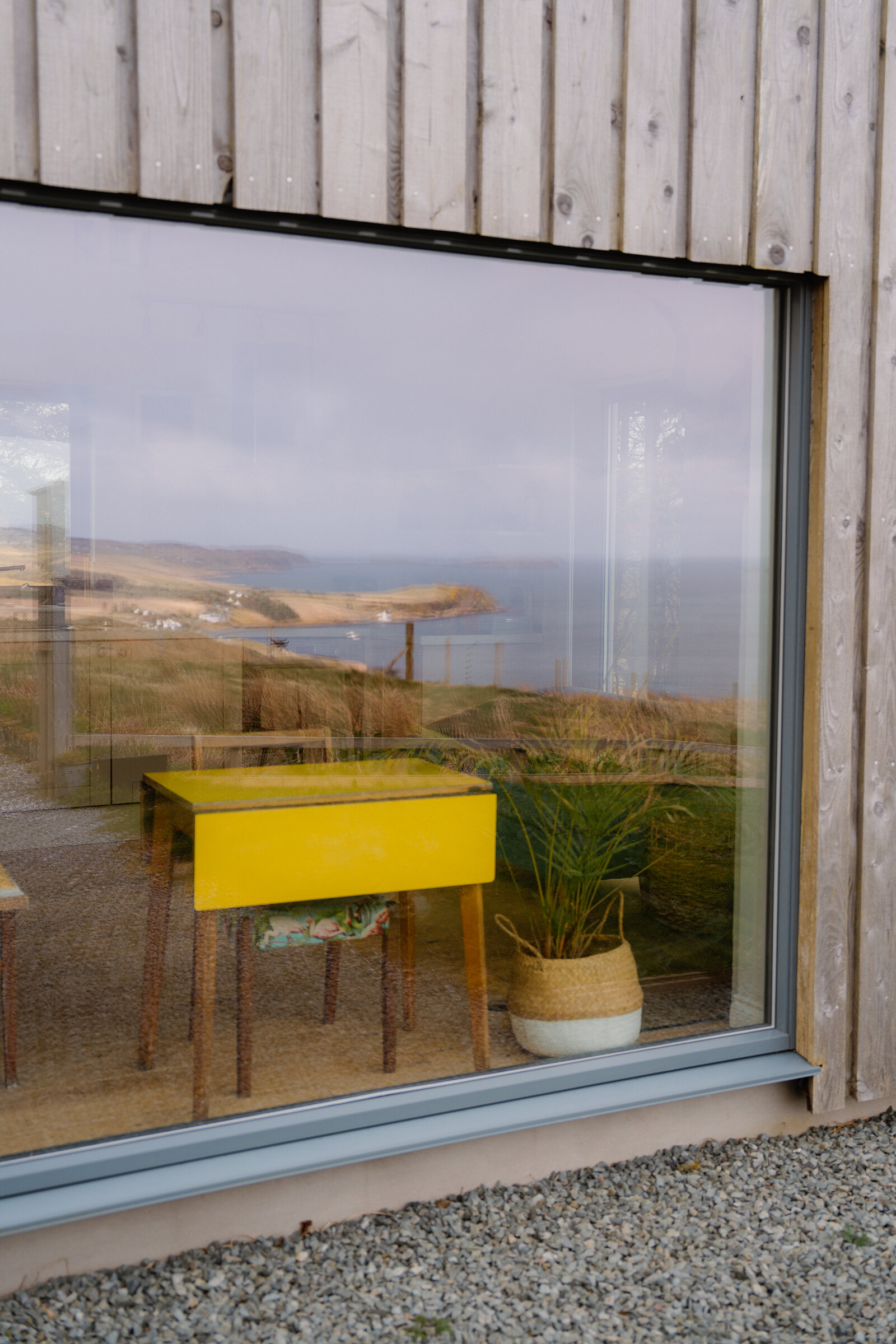 A yellow desk and woven basket sit beside a large window overlooking a coastal headland.