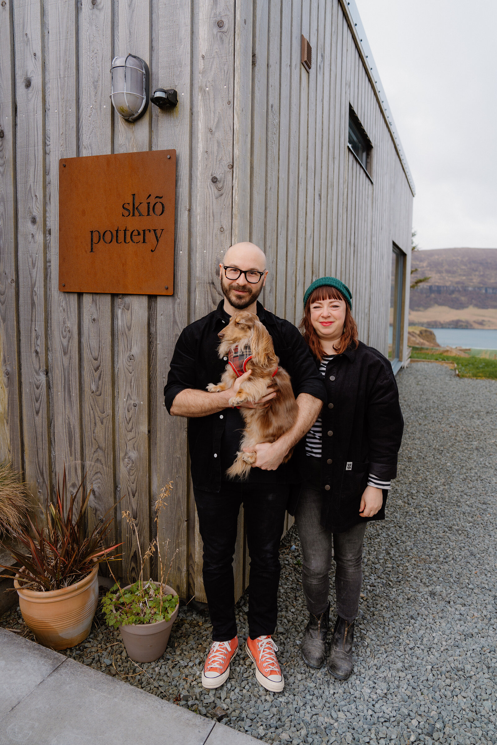 A man holding a small dog stands beside a woman before a sign reading 'skíõ pottery' on a timber-clad building.