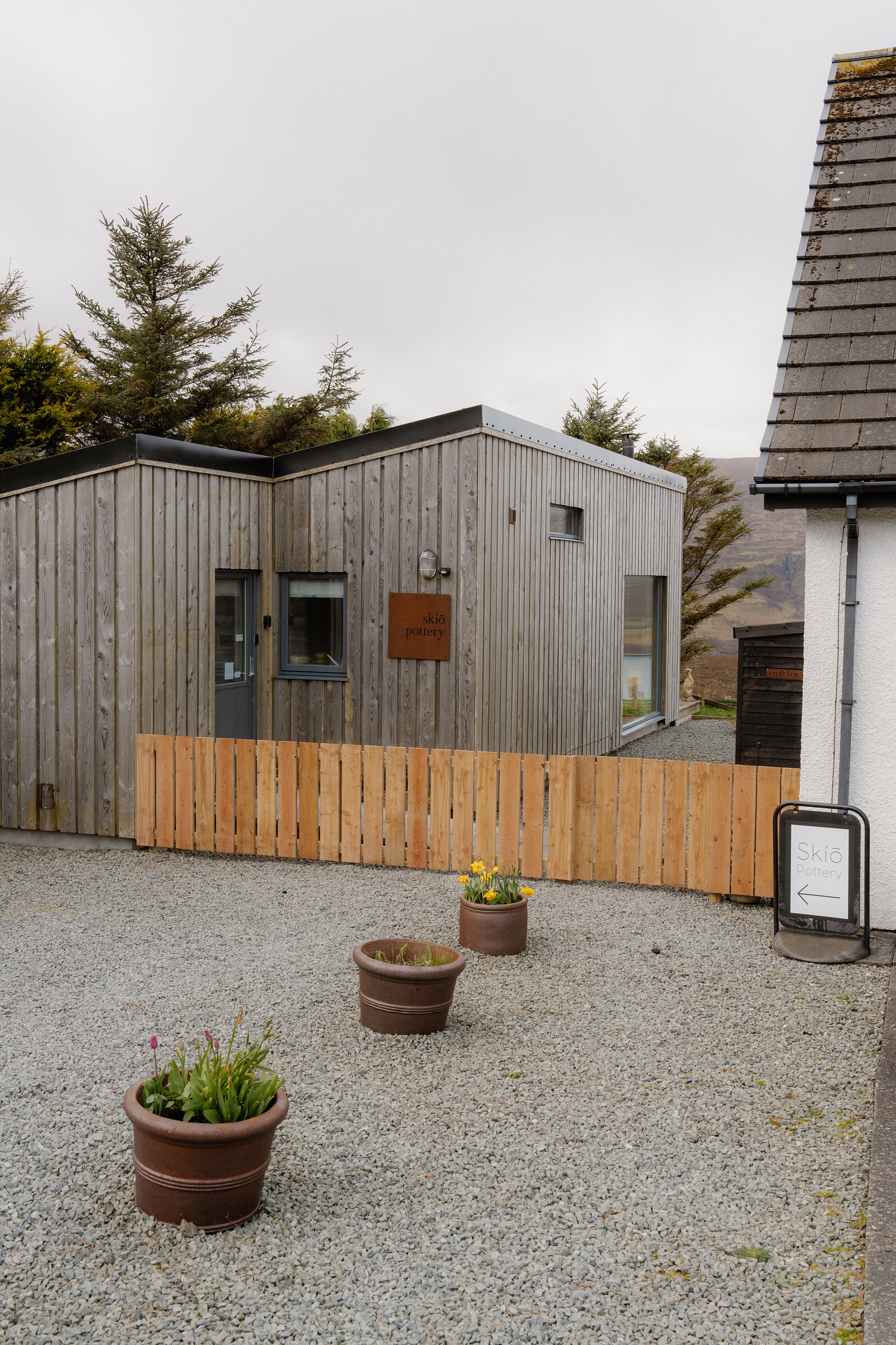 A timber-clad building with a 'Skíõ' directional sign sits behind a gravel forecourt and potted plants.