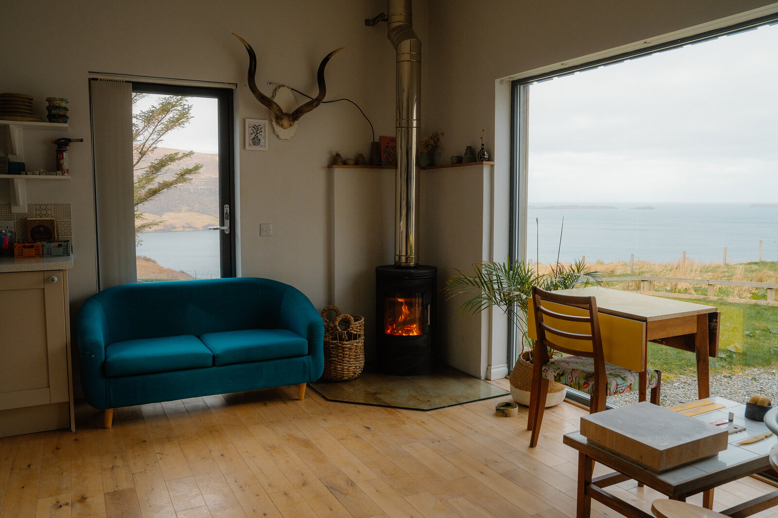 A teal sofa and wood-burning stove sit before an open sliding door overlooking the sea.