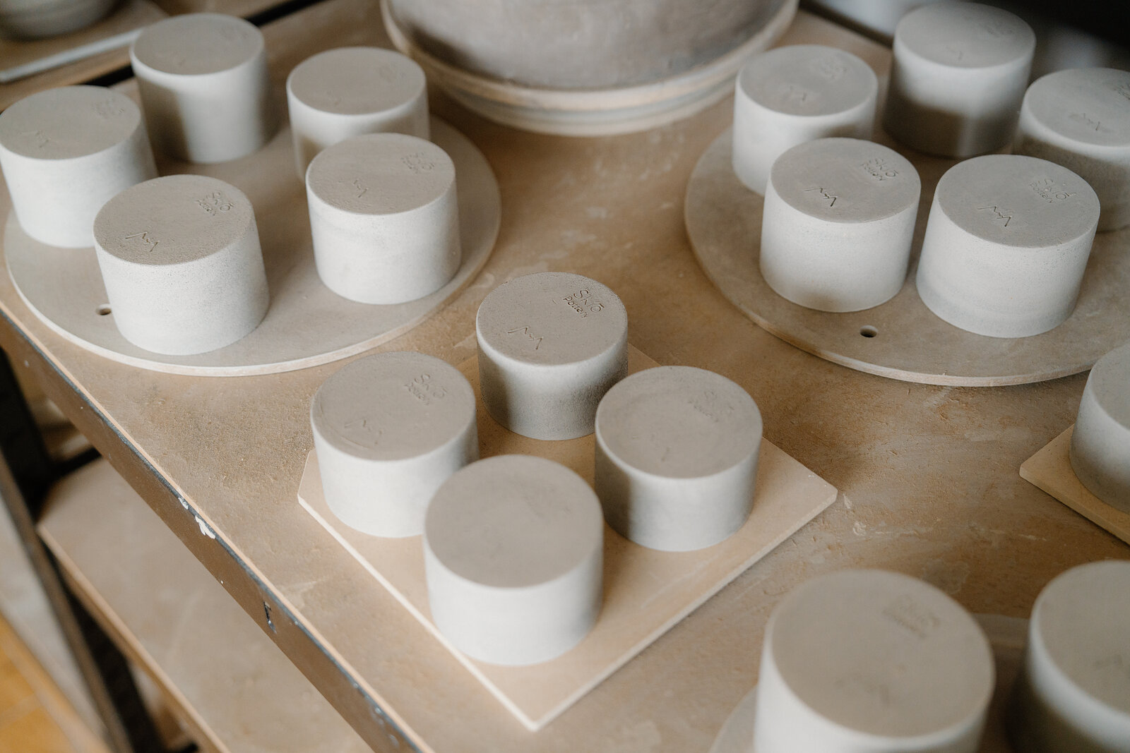 Rows of cylindrical clay blanks sit drying on wooden bats in a pottery studio.