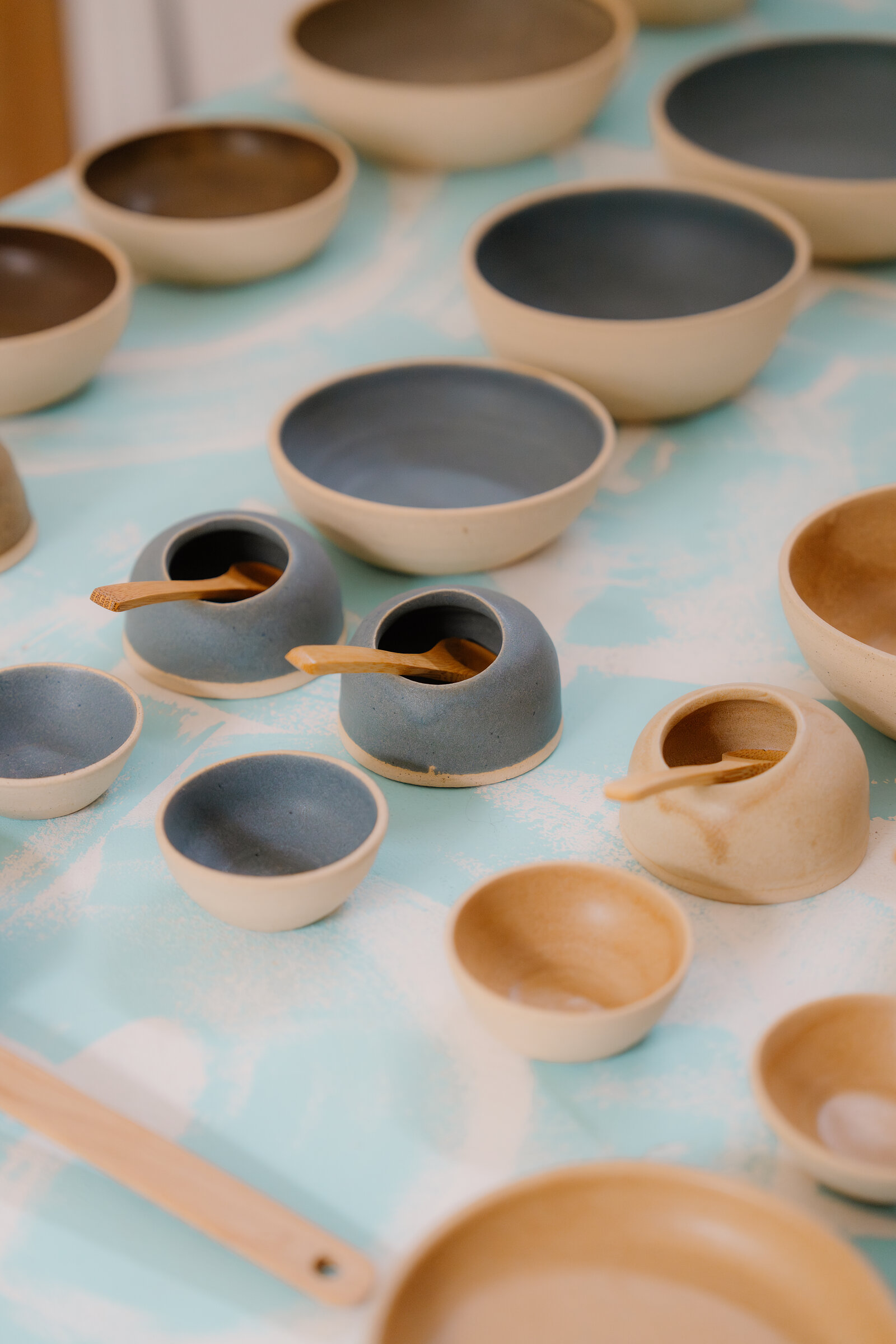 Glazed stoneware bowls and small lidded pots with wooden spoons are arranged on a patterned cloth.
