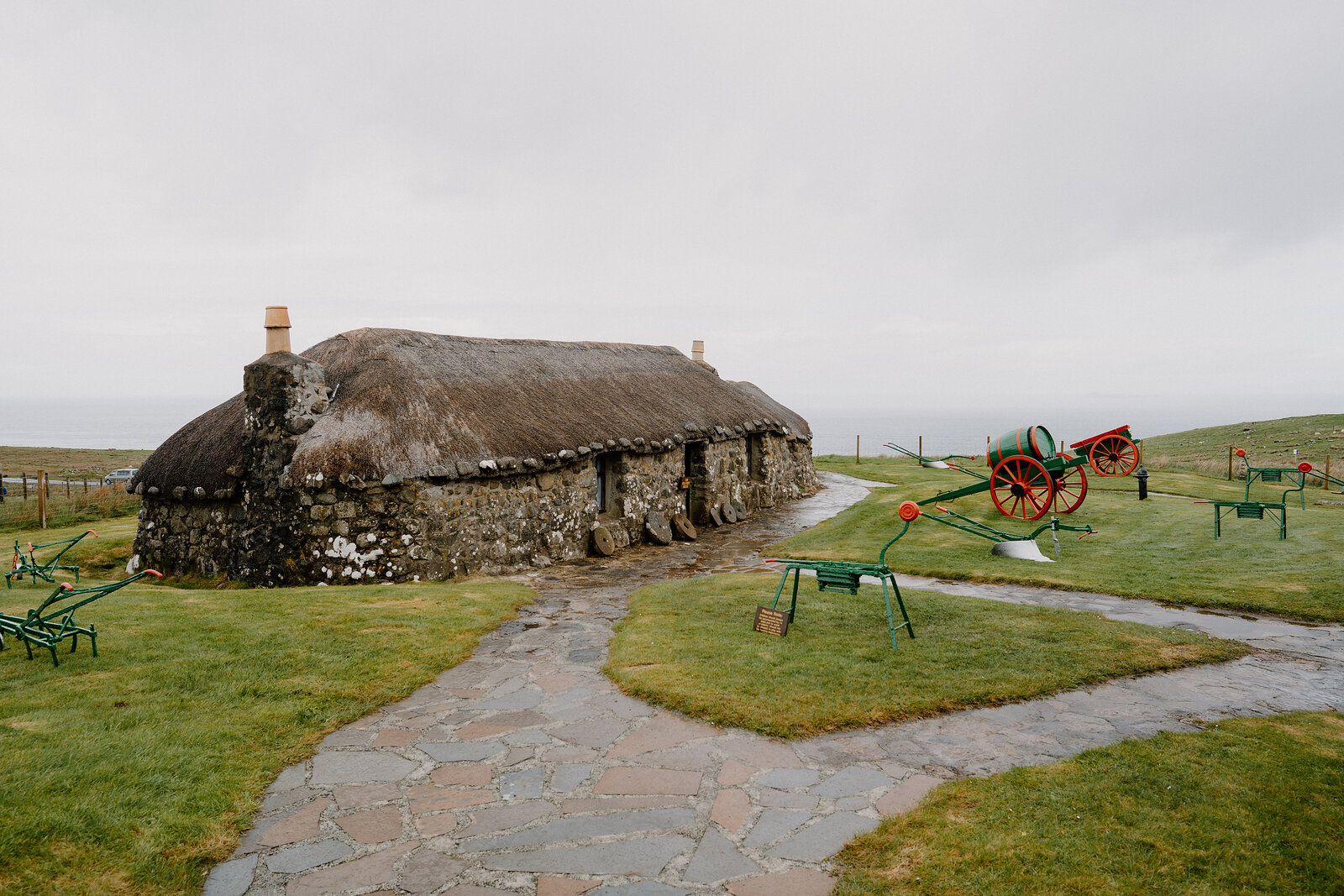 A thatched blackhouse sits beside a stone path in a grassy museum courtyard, farm carts in the background.