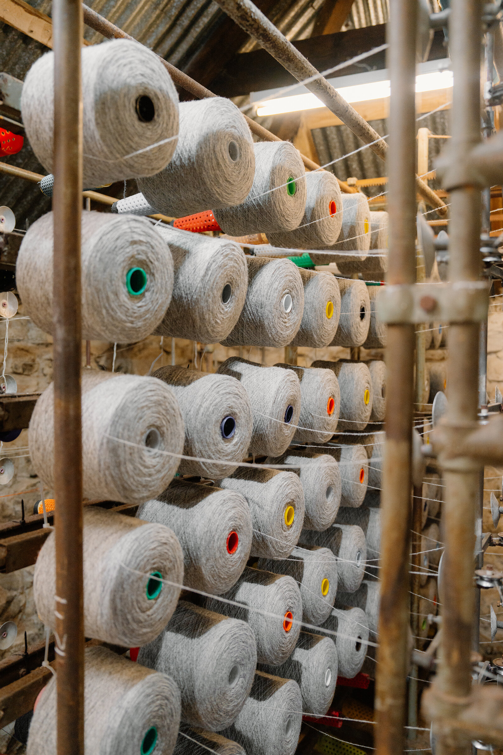 Grey yarn bobbins with coloured tips are stacked in rows on a wooden rack.