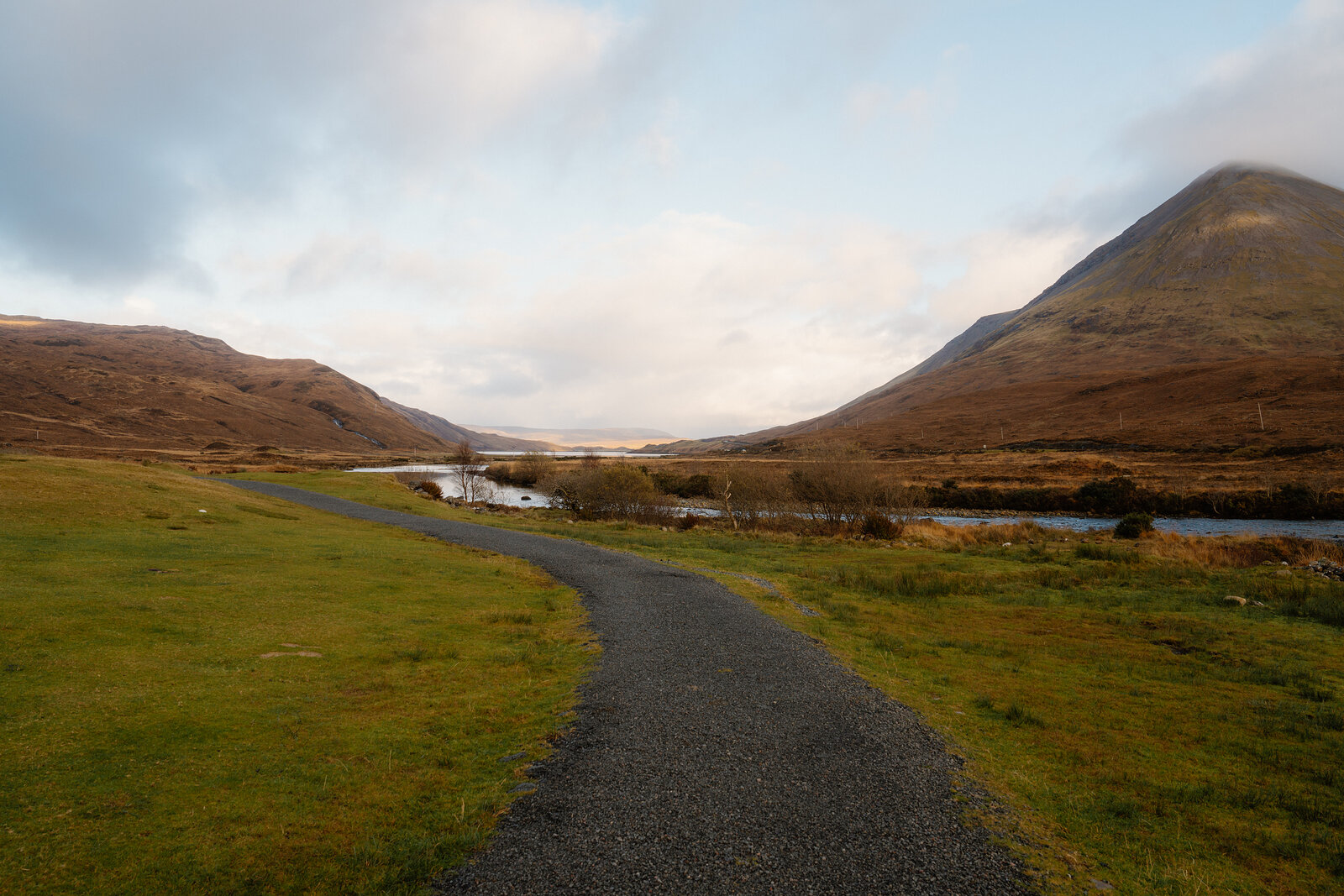 A gravel path curves beside a river toward distant mountains under a cloudy sky.