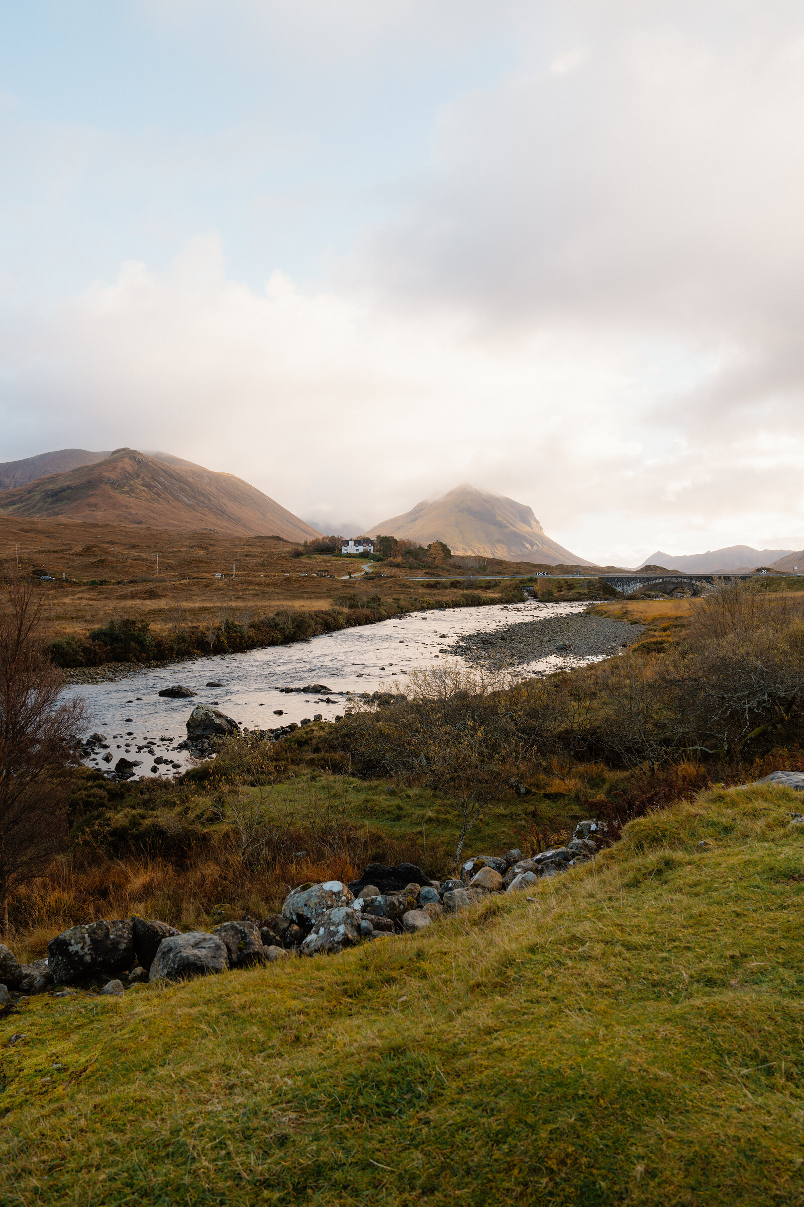 A river curves past a white house near a stone bridge, mountains rising beyond in soft light.