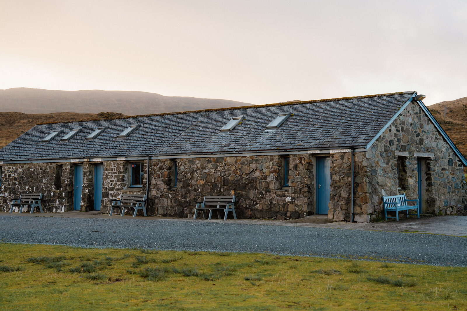 A long stone cottage with blue doors and benches sits before rolling hills.